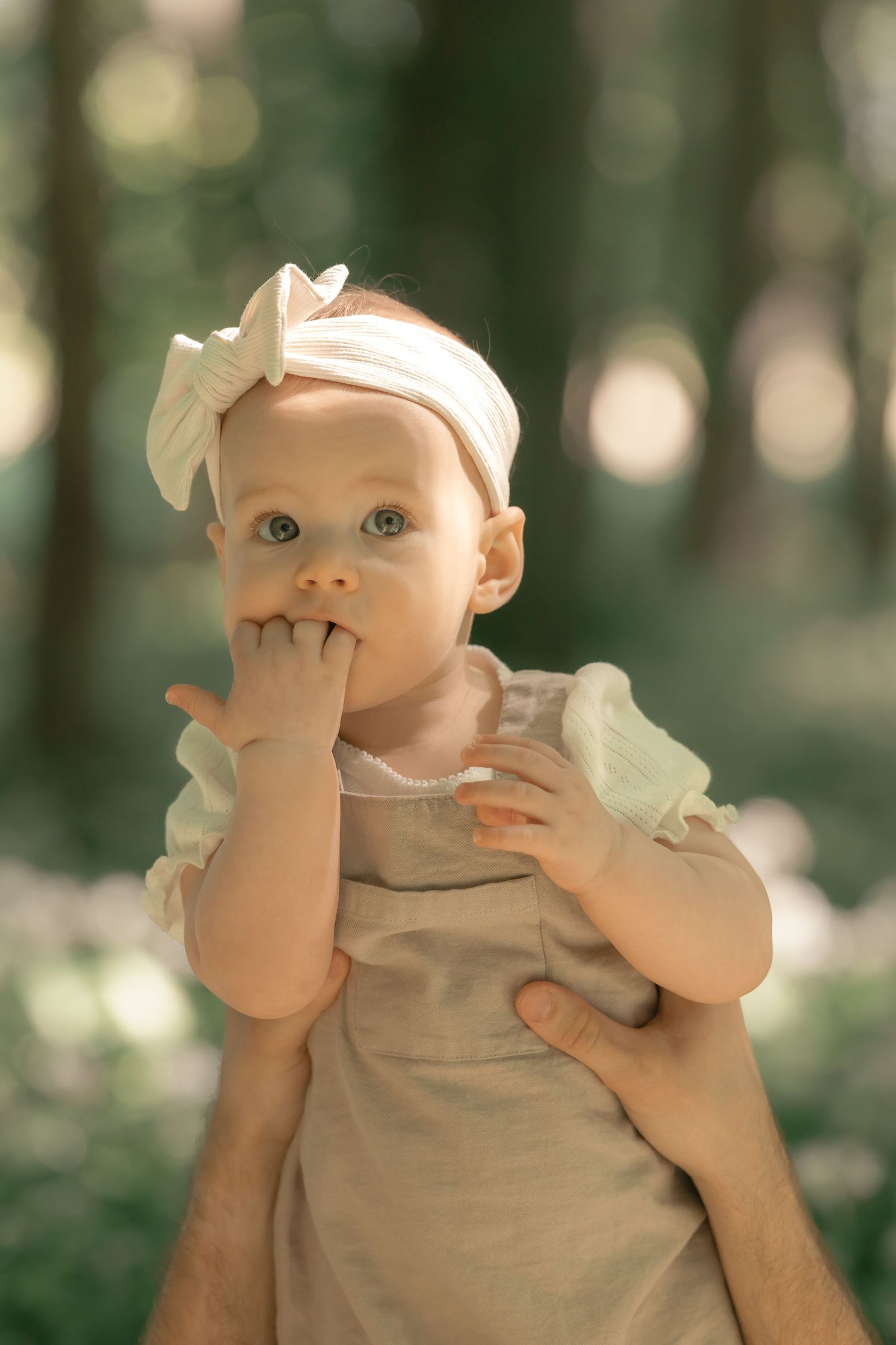 Cute baby girl with white bow headband putting finger in mouth while being held outdoors.