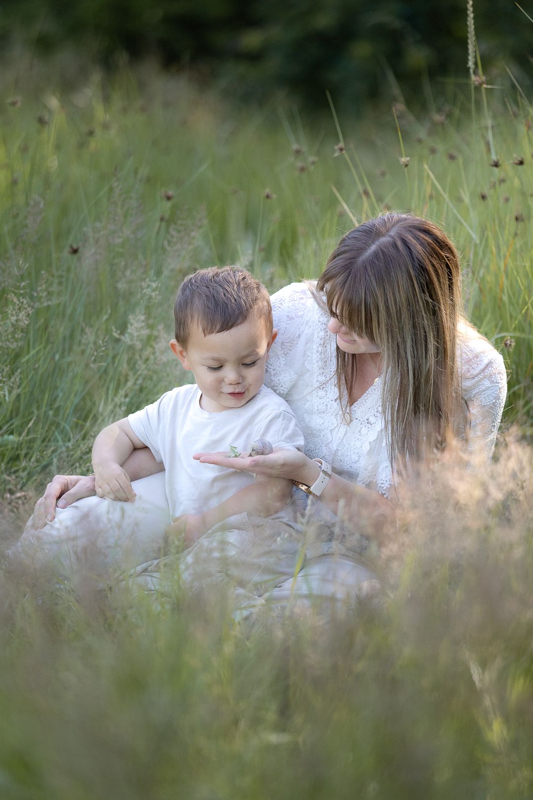 Mother and toddler son sitting together in tall green grass in a meadow.