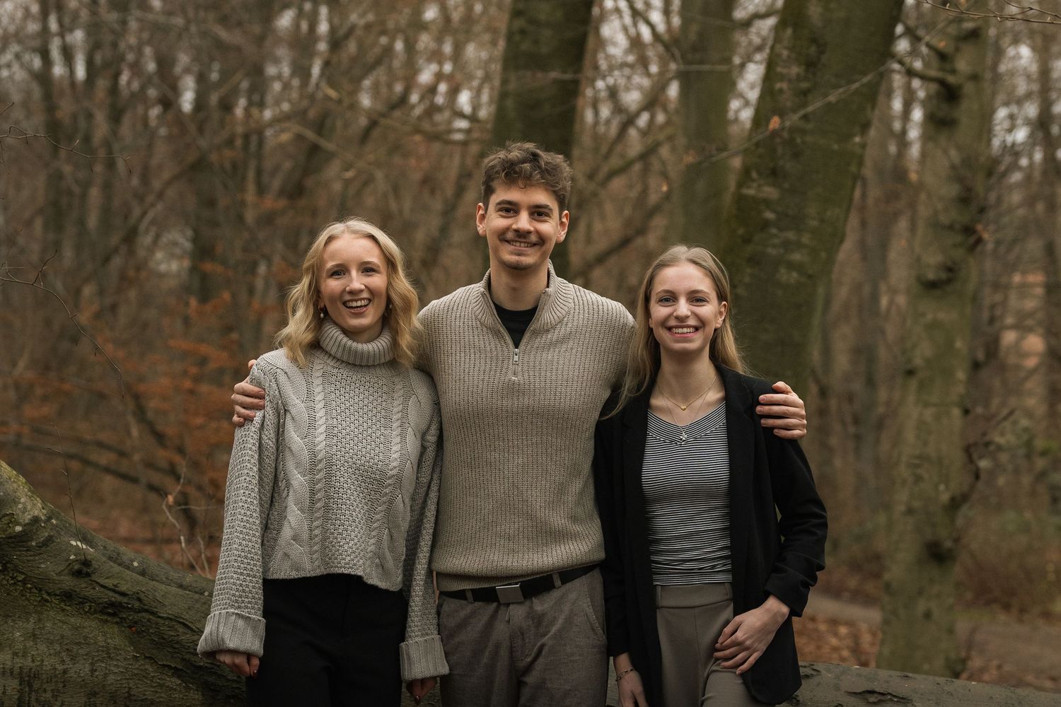 Three young adults smile together in an autumn forest, posing near a fallen log.