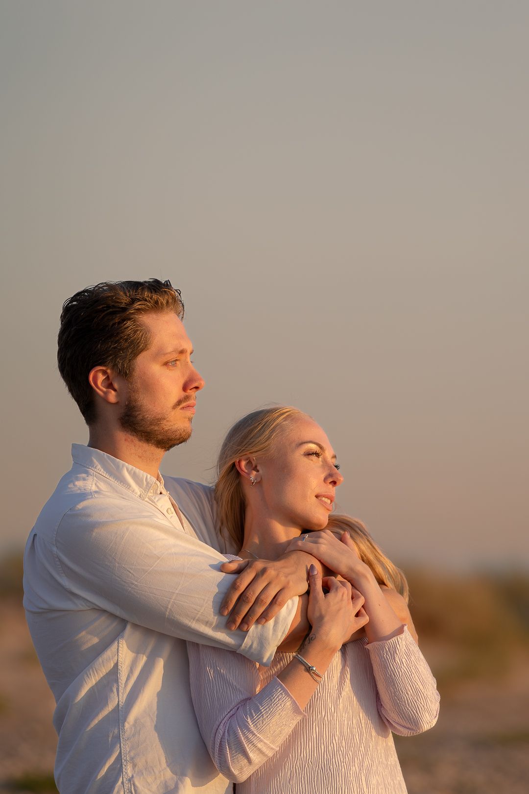Couple embracing at sunset on rocky beach, man in white shirt hugging woman from behind.