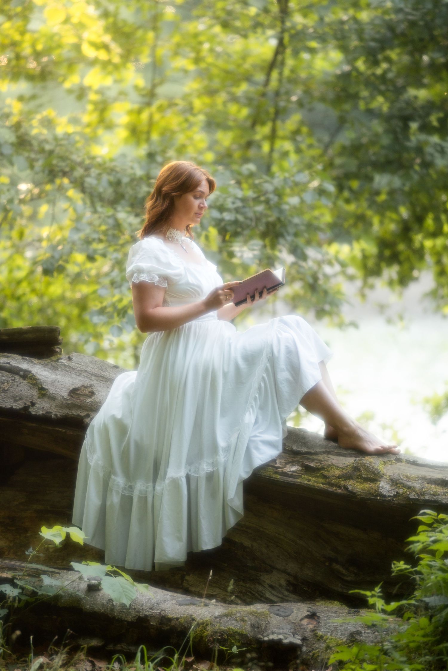 Woman in white dress reading a book while sitting on a log in a lush green forest setting.
