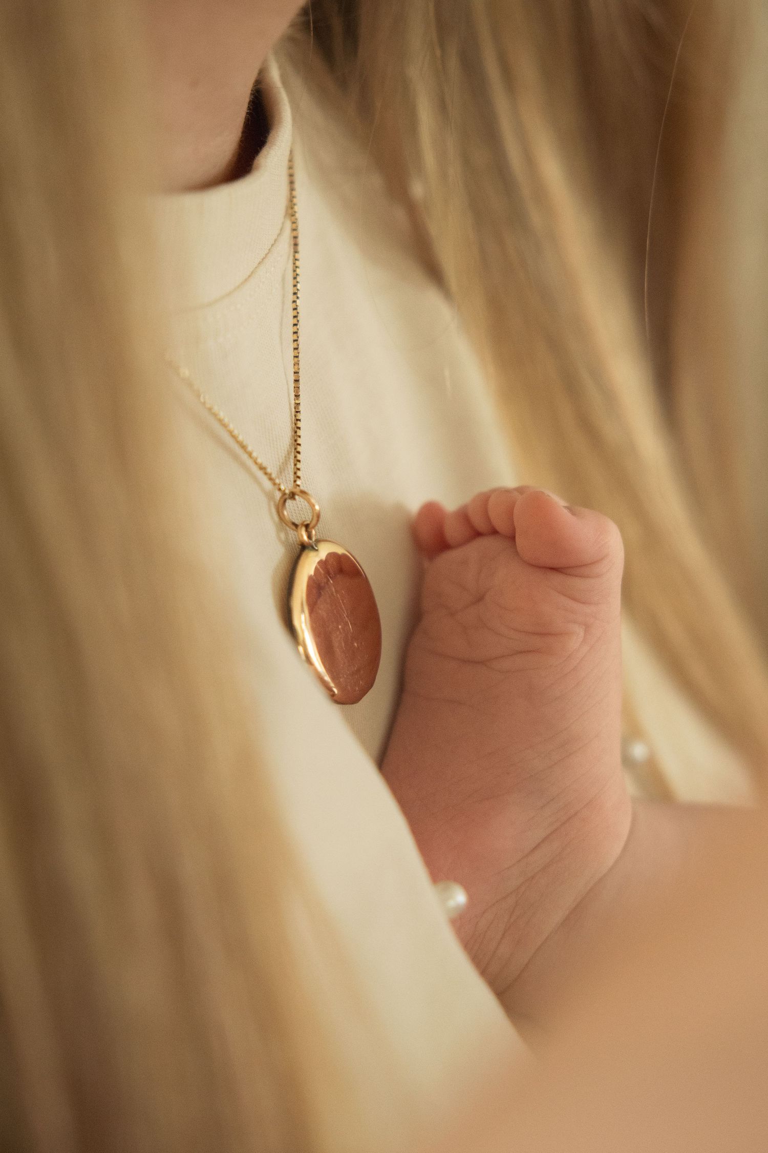 Close-up of a newborn baby foot resting against a mother's chest beside a gold oval locket necklace.