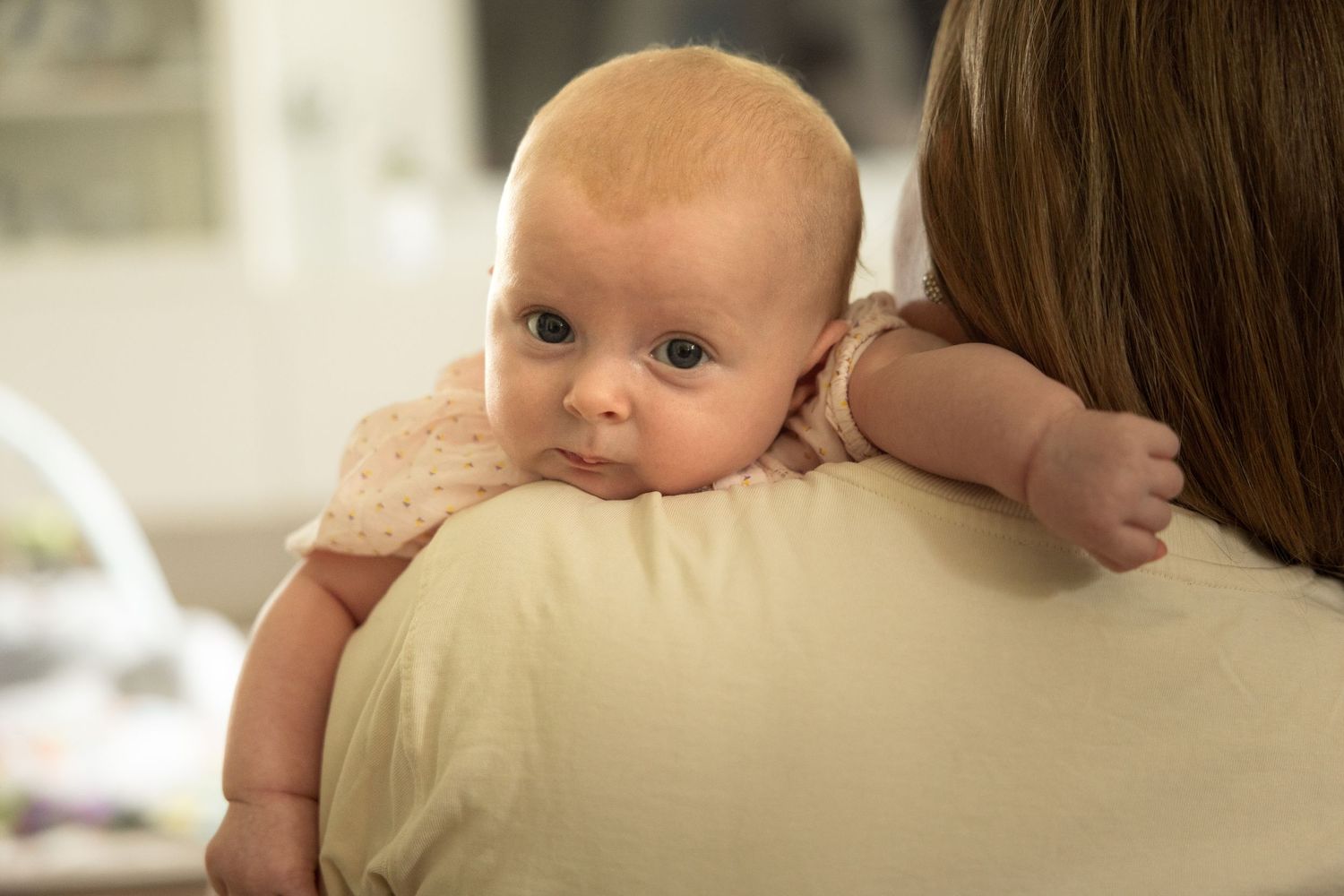 Newborn baby resting on mother's shoulder, looking back at camera with curious blue eyes.