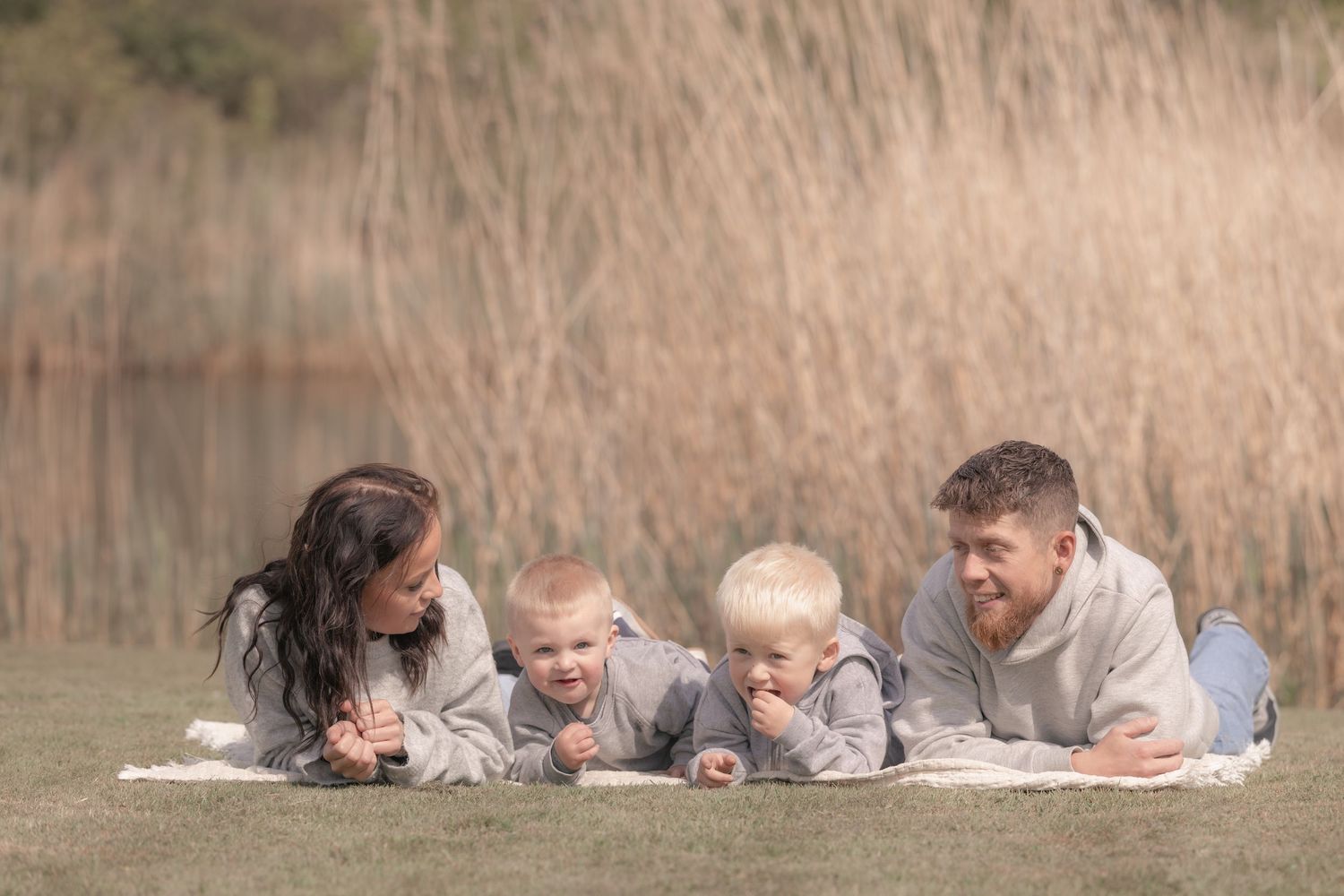 Family of four lying on a blanket on grass, smiling and laughing together near tall reeds.