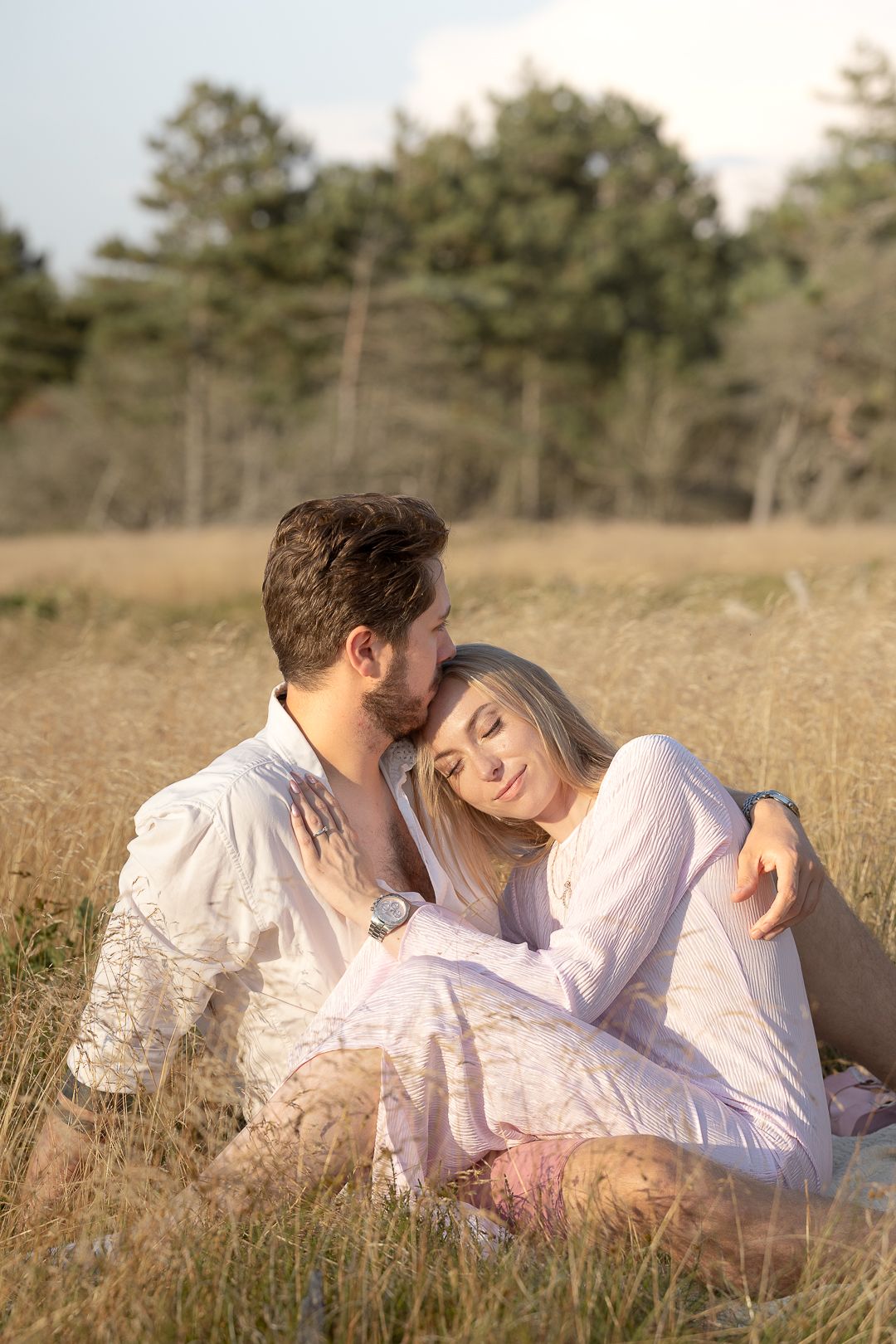 Romantic couple sitting in golden field, woman in pink dress resting on man's shoulder, surrounded by trees.