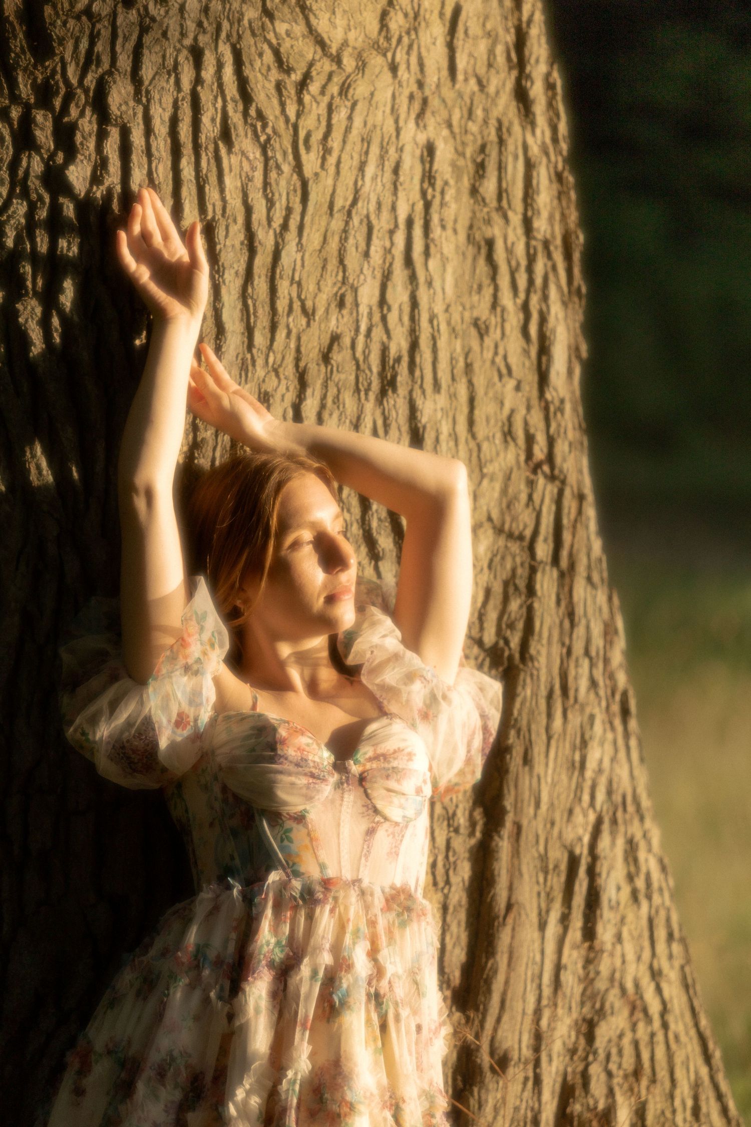 Woman in floral dress leaning against large tree trunk in golden sunlight with arms raised gracefully.