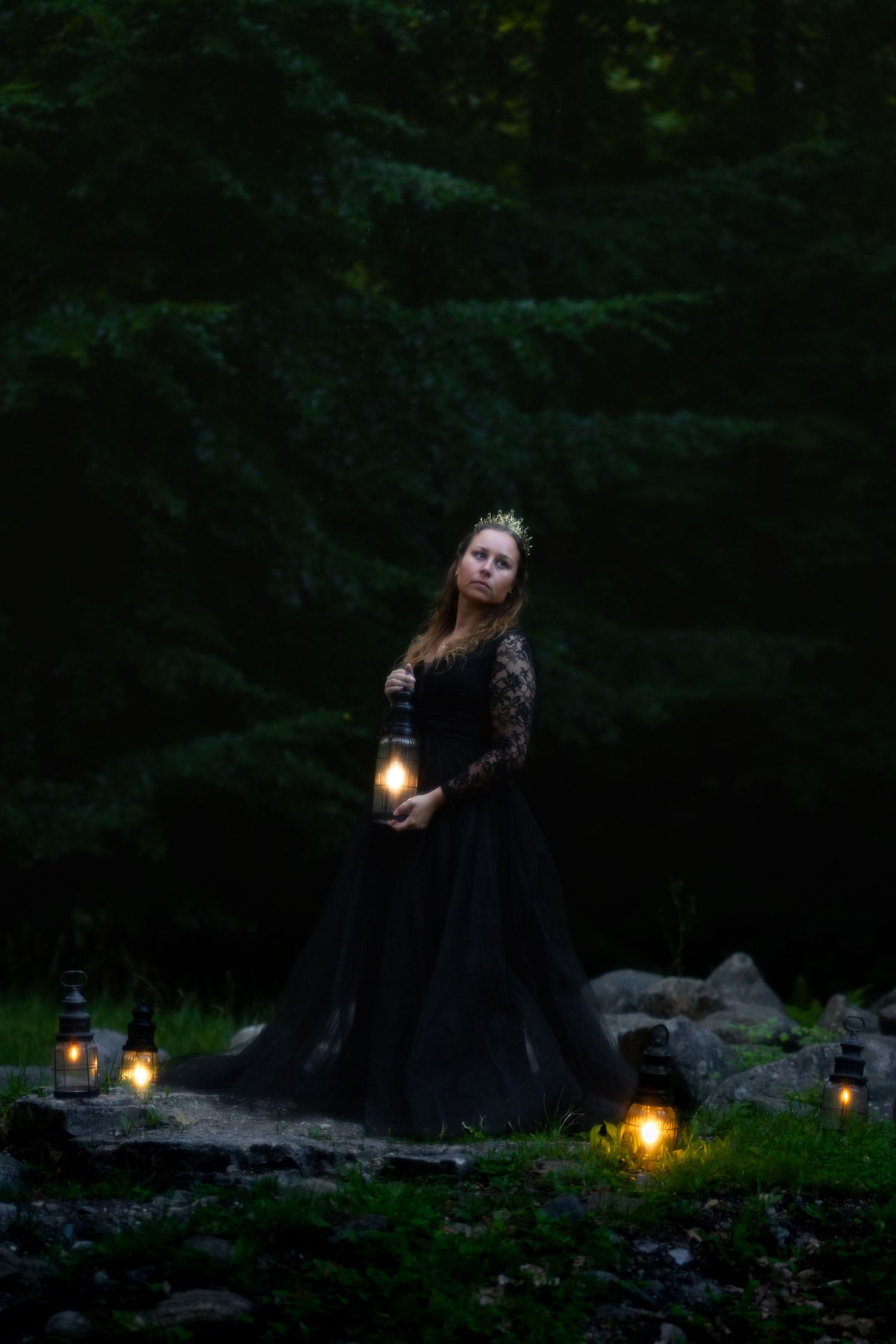 Woman in black lace gown holding lantern in dark forest with glowing lights on rocks at dusk.
