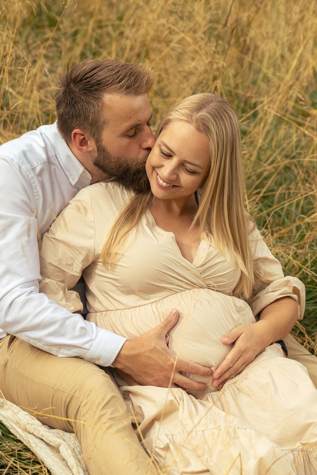 Couple sitting in tall grass, man kissing pregnant woman's cheek while holding her bump.