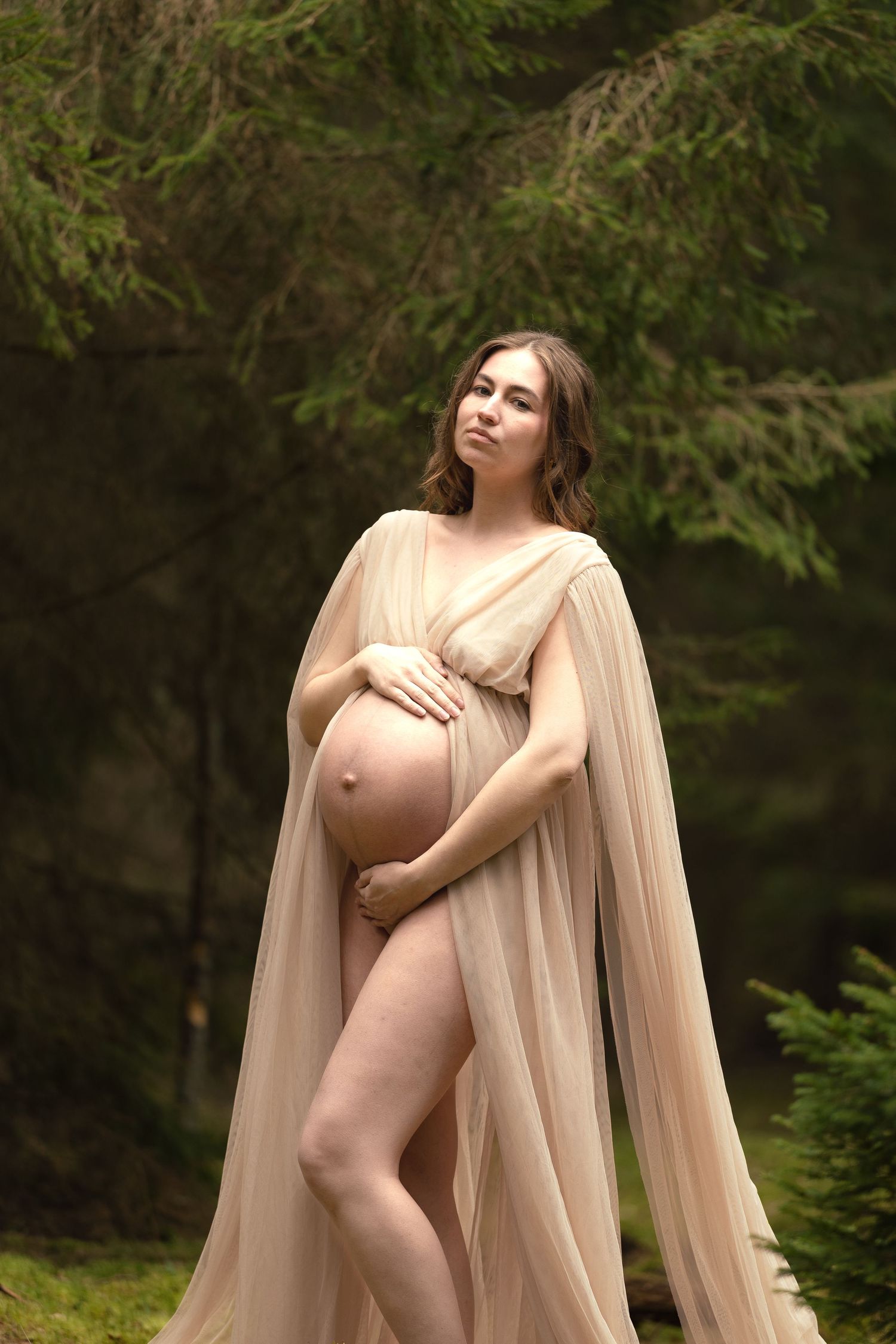 Pregnant woman in flowing beige sheer cape gown posing among green pine trees outdoors.