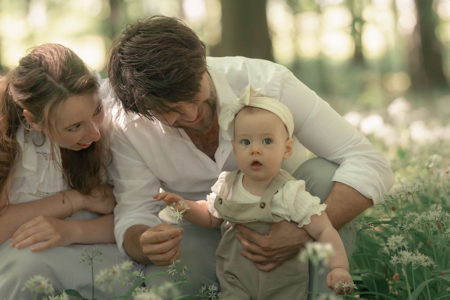 Young parents sitting in wildflower meadow with curious baby in white forest setting.