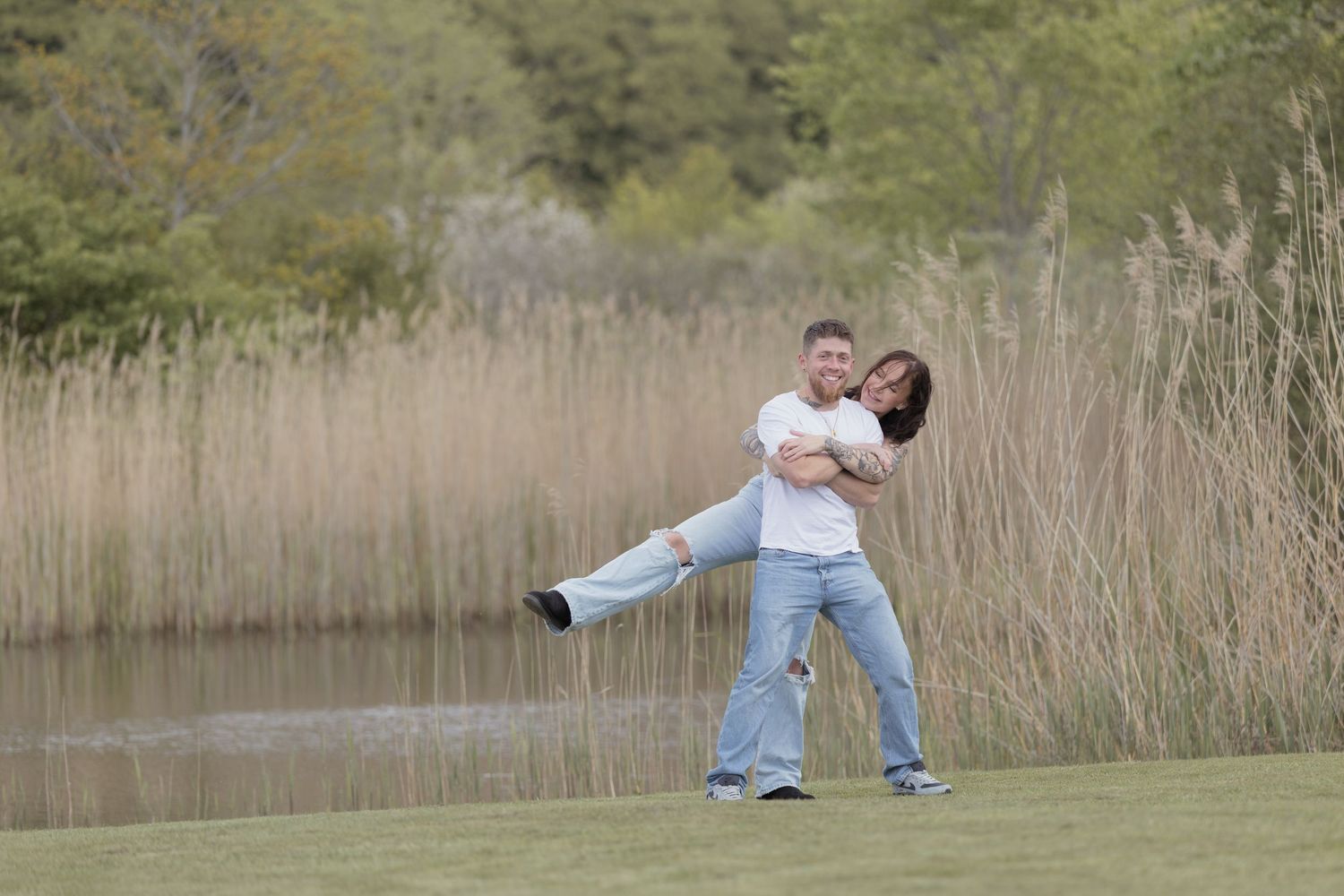 Man lifting and spinning woman in his arms near a serene pond with tall reeds.