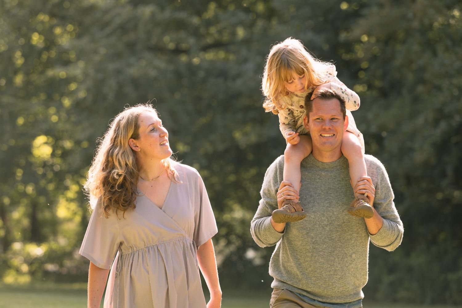Happy family walking outdoors, father carrying child on shoulders, mother smiling, lush green trees in background.
