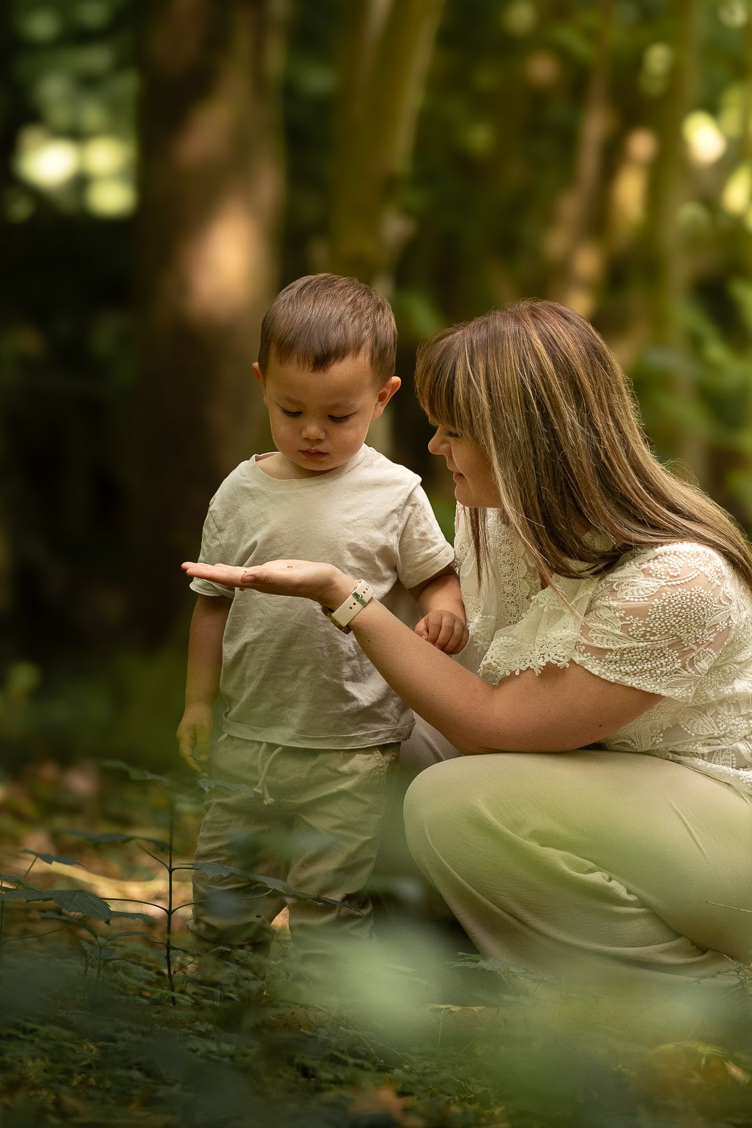 Mother crouching beside toddler son exploring nature in a lush green forest.