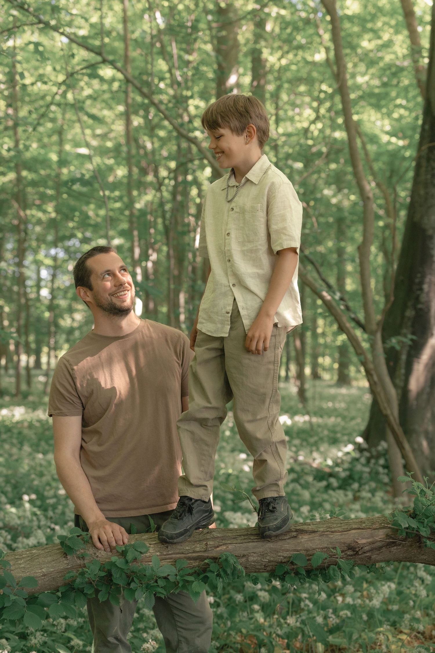 Father watching son standing on fallen log in sunlit green forest clearing.