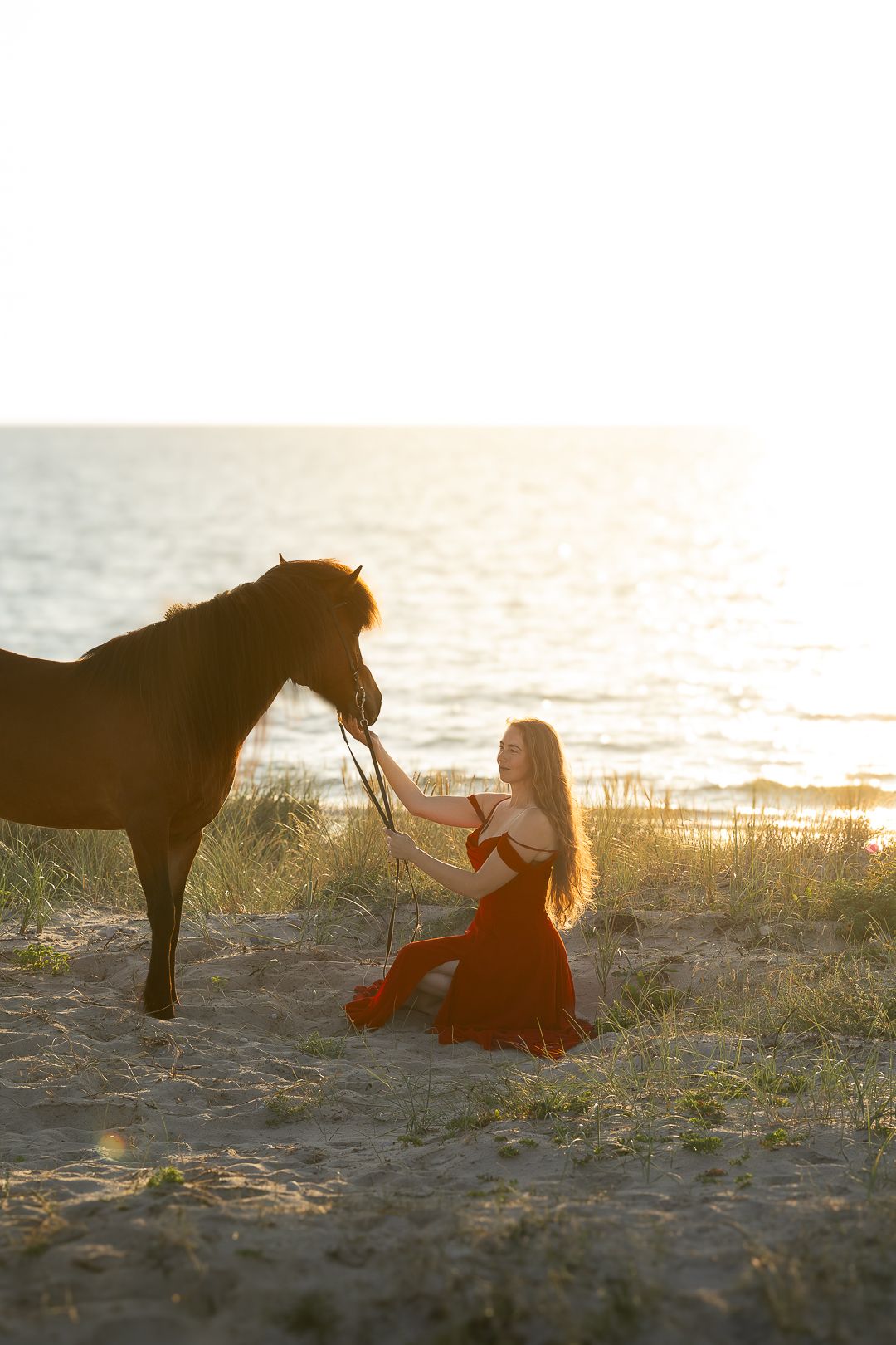 Woman in red dress kneeling on sandy beach holding lead rope of brown horse at sunset.