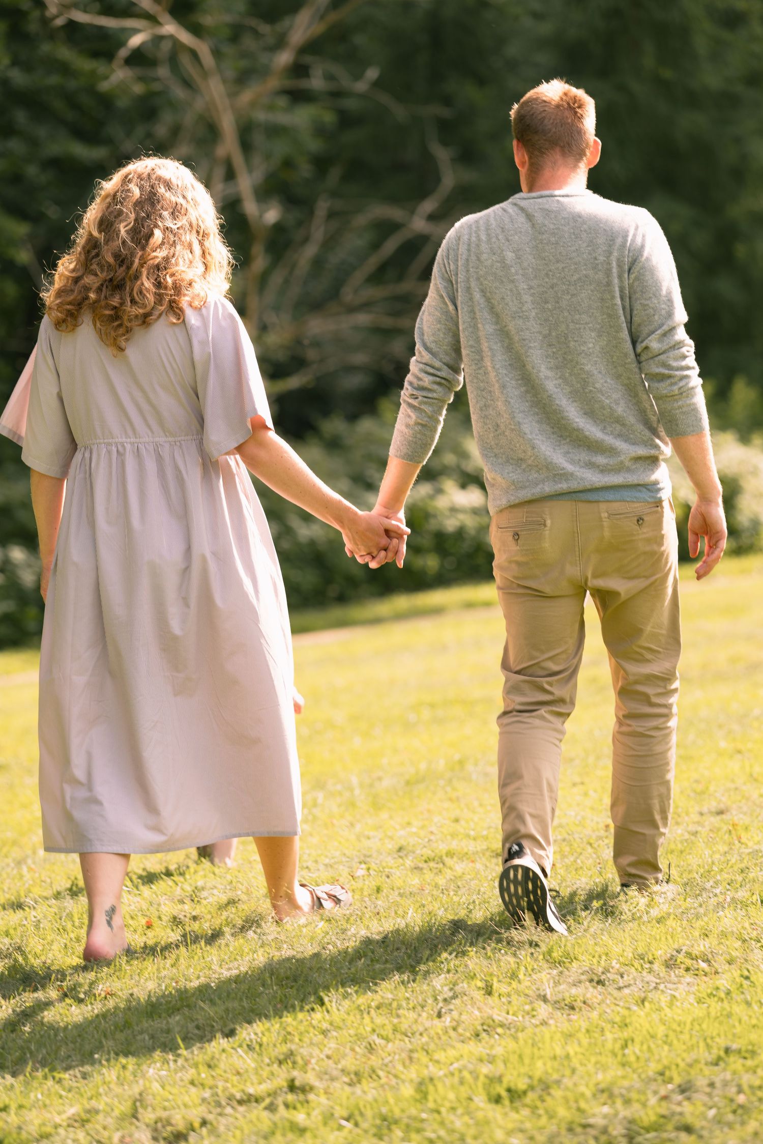 Couple holding hands walking through lush green park, viewed from behind, woman in floral dress.