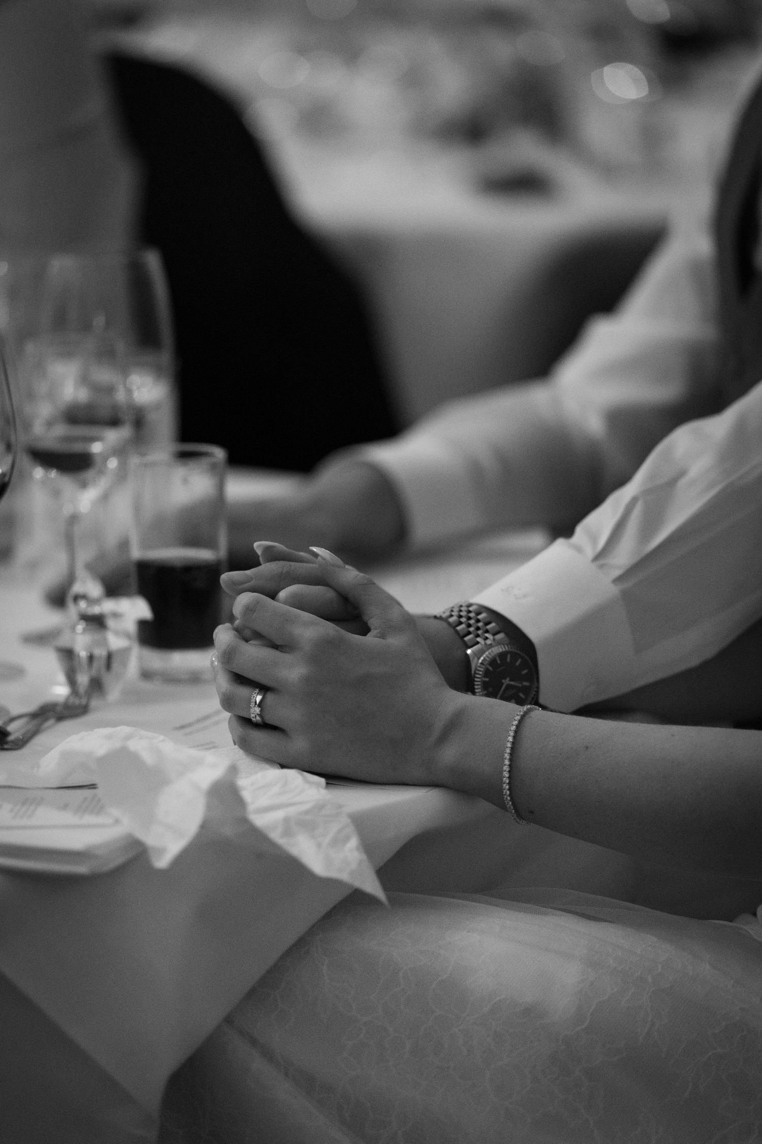 Close-up black and white photo of a wedding guest's hands clasped on a white tablecloth with wine glasses nearby.