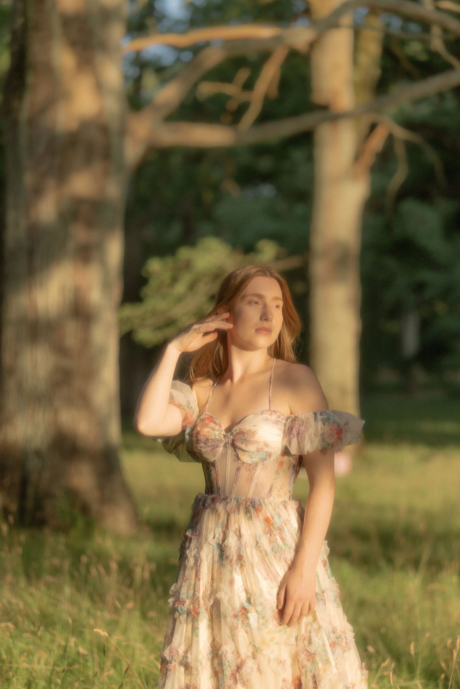 Woman in floral off-shoulder dress standing in sunlit forest clearing, golden hour light.