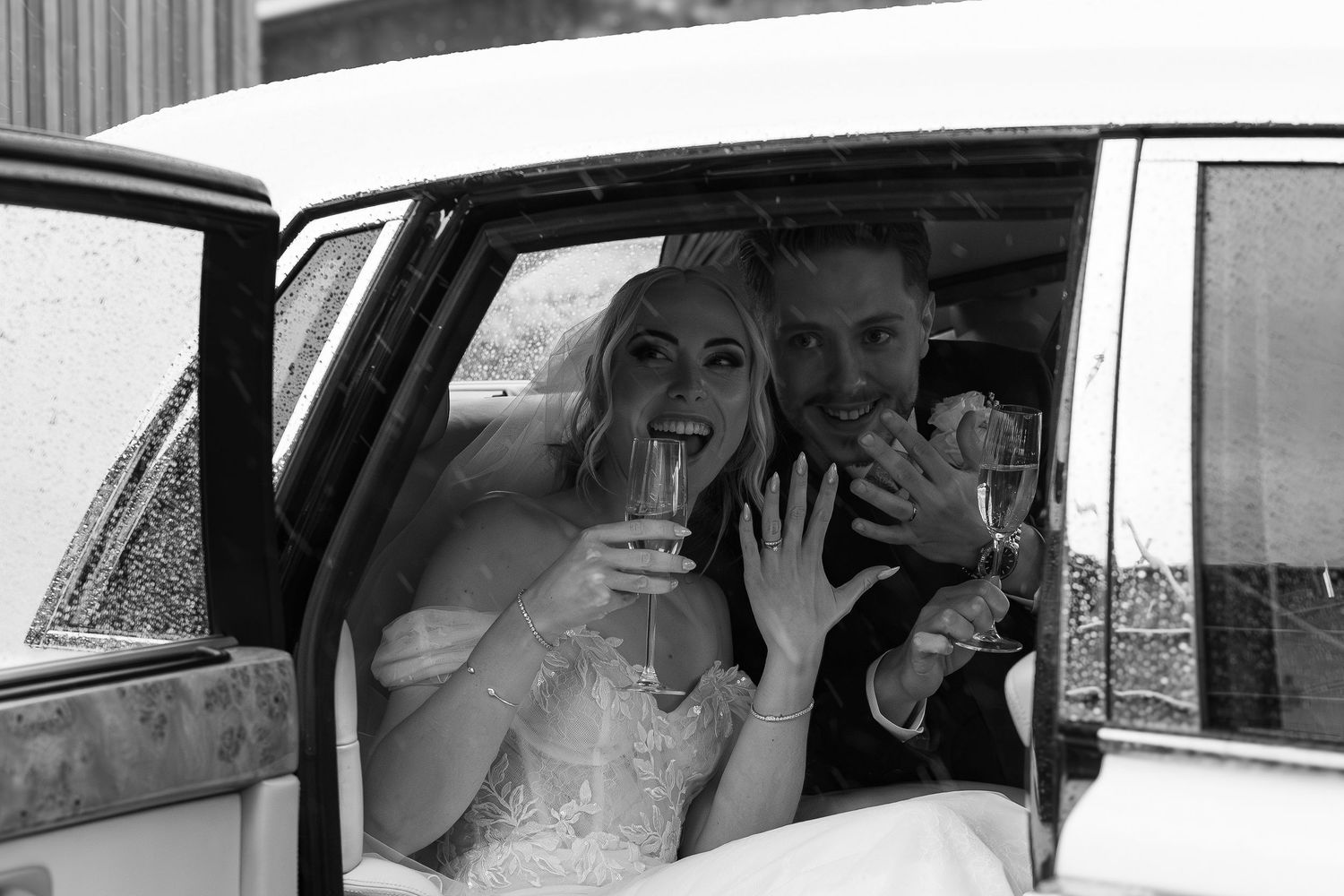 Newlyweds celebrate in vintage wedding car, bride holding champagne and showing ring in black and white.