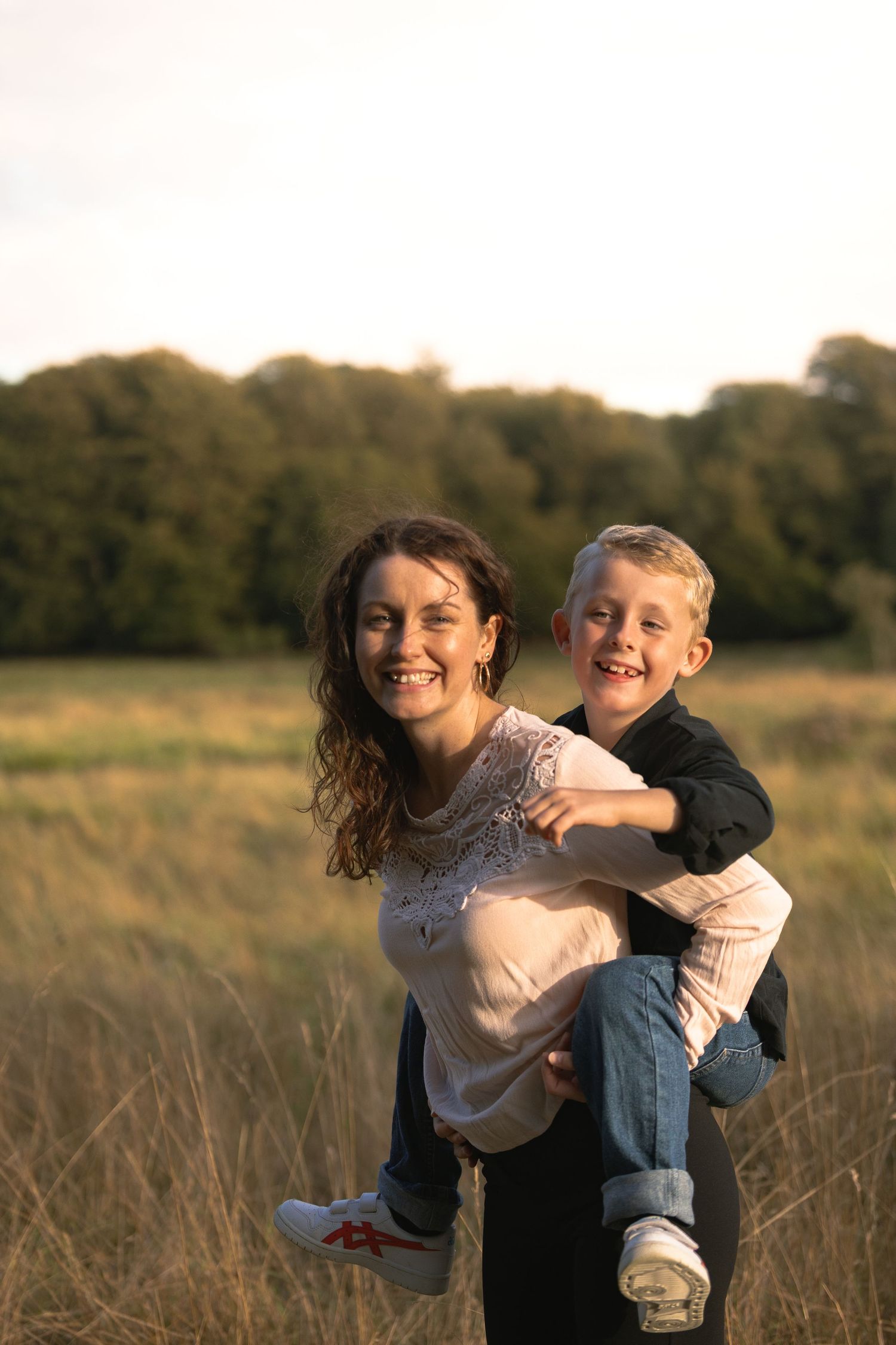 Happy mother giving smiling young son a piggyback ride in golden sunlit meadow field.