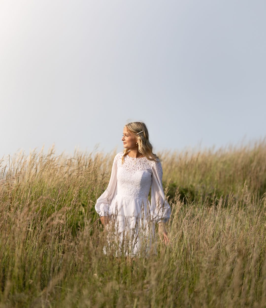 Blonde woman in white boho lace dress standing in tall golden grass field under clear sky.