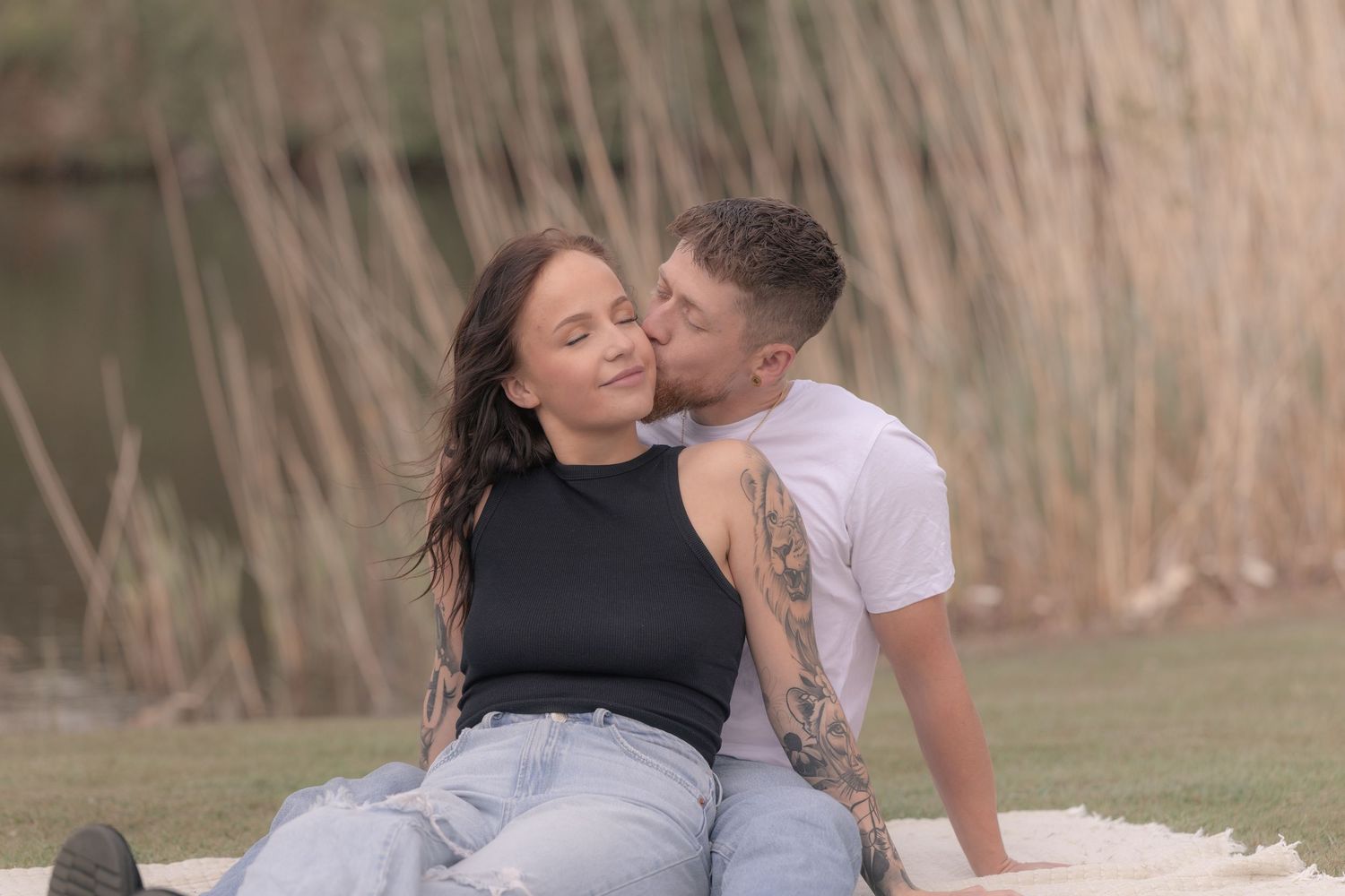 Couple sitting on a blanket near tall reeds as man gently kisses woman on the cheek.