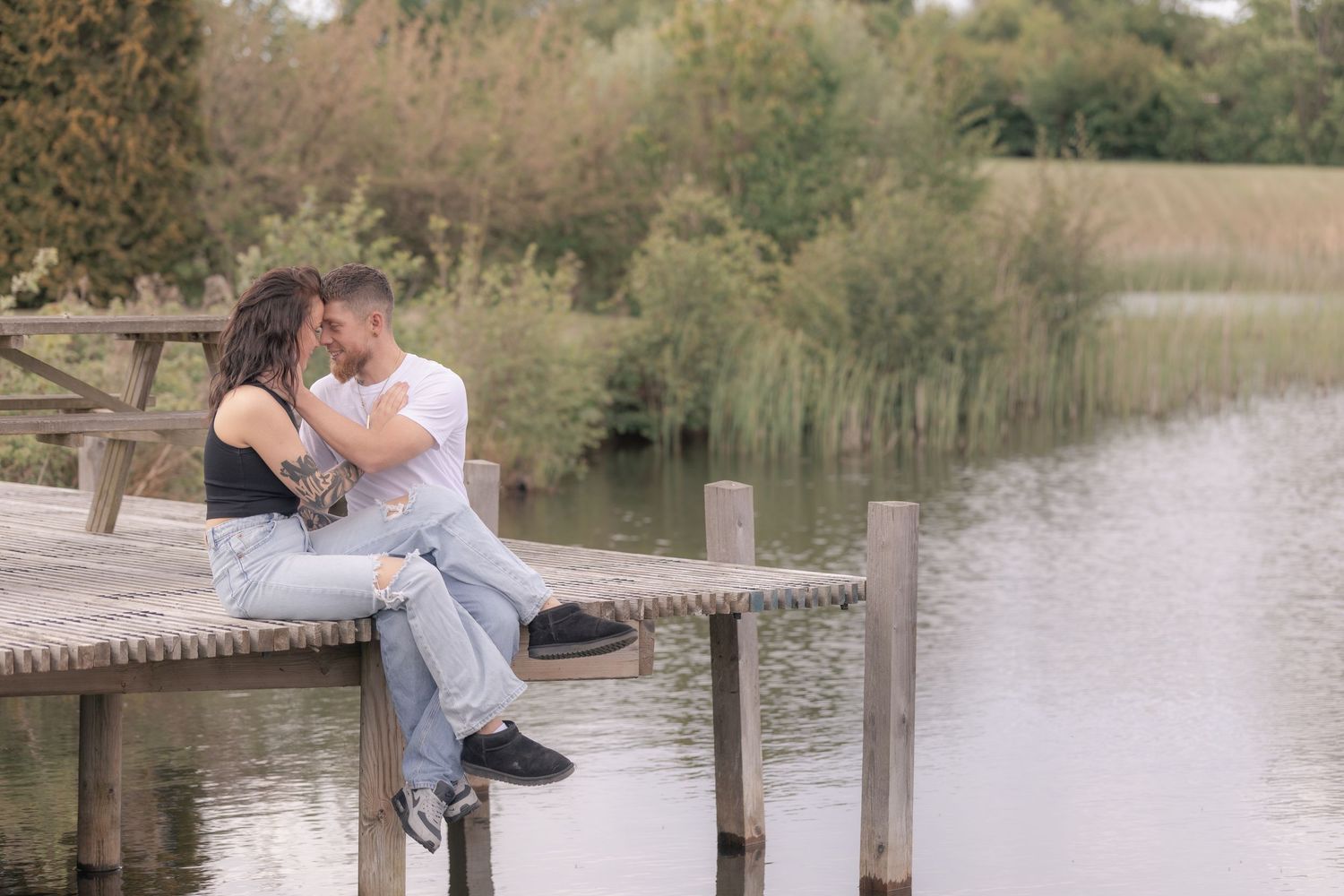 Couple sitting on a wooden dock by a lake, leaning in for a kiss surrounded by lush greenery.