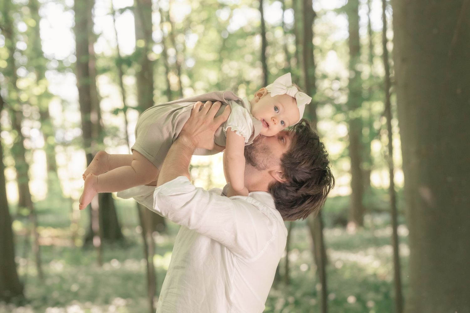 A father lifts his laughing baby girl high in the air in a sunlit woodland setting.