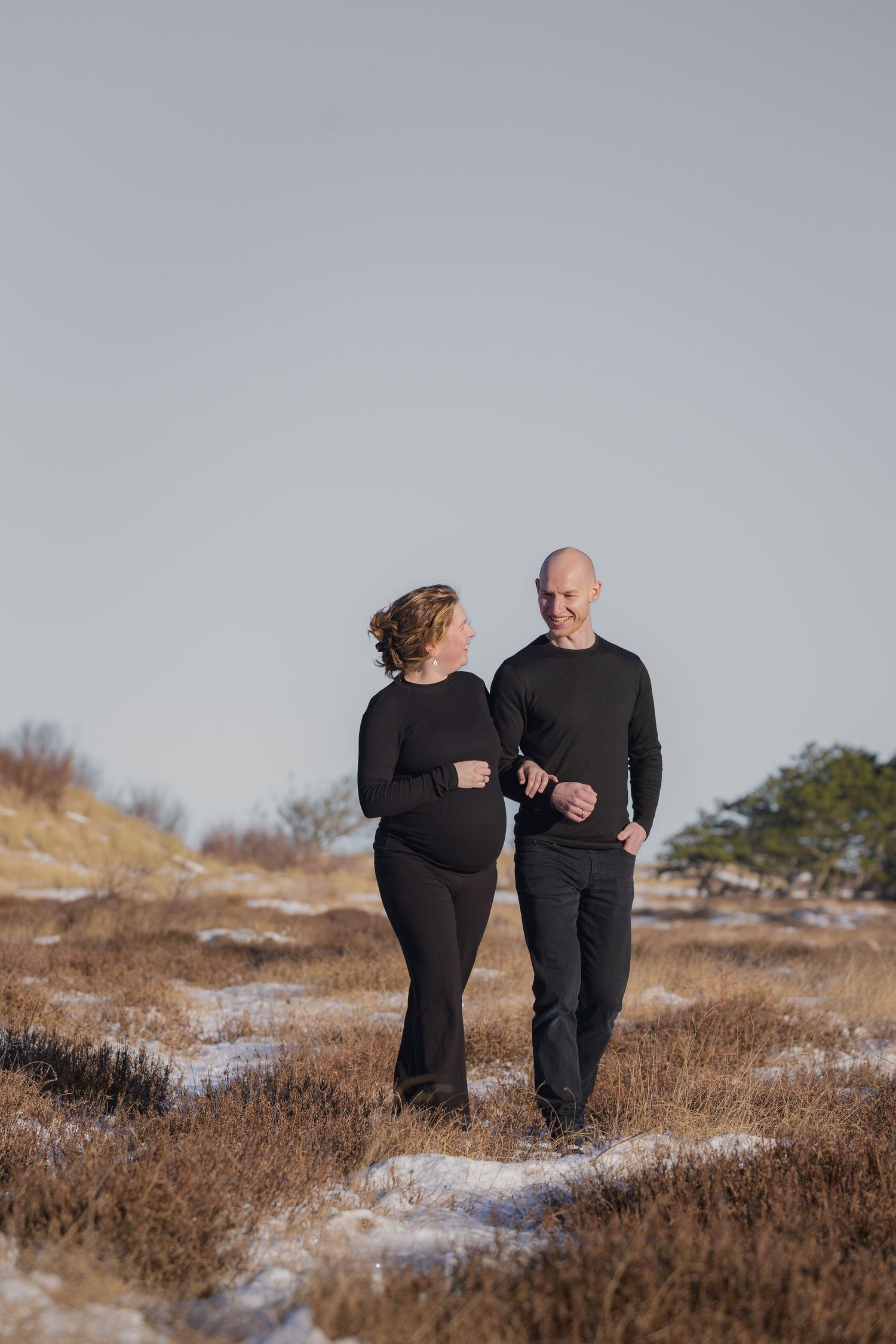 Happy pregnant couple dressed in black walking together through a snowy winter heathland.