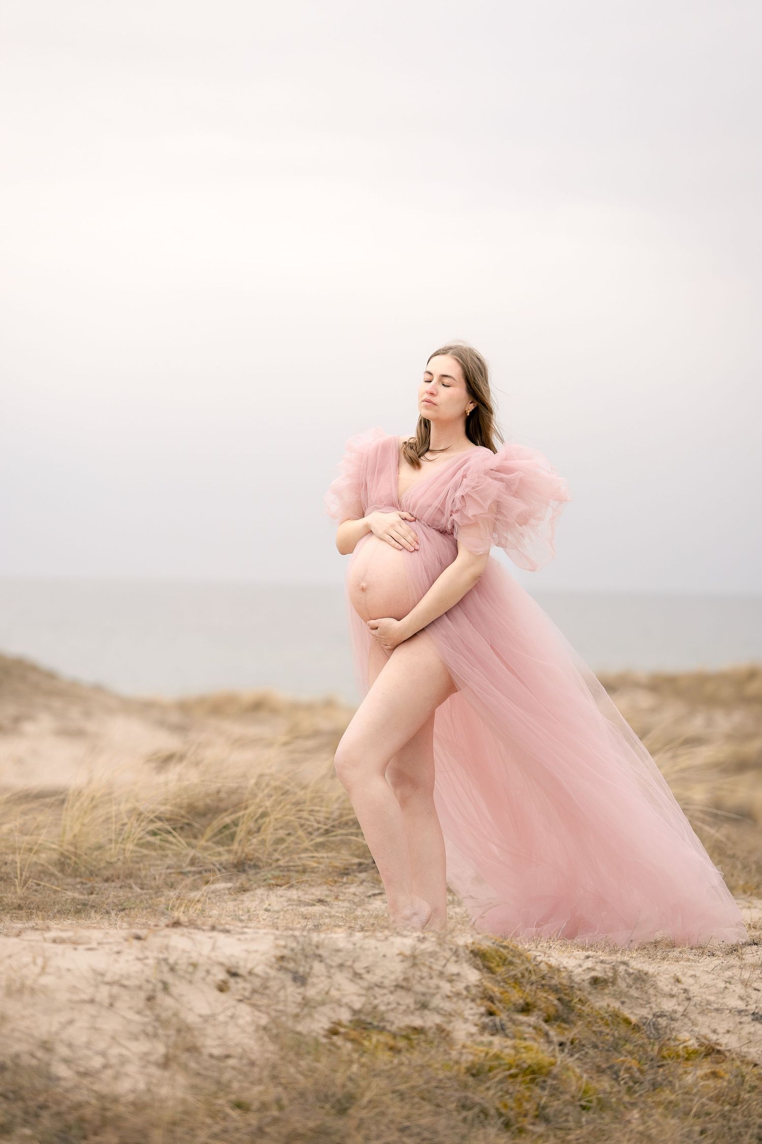 Pregnant woman in flowing pink tulle gown cradling her baby bump on sandy beach dunes with ocean backdrop.
