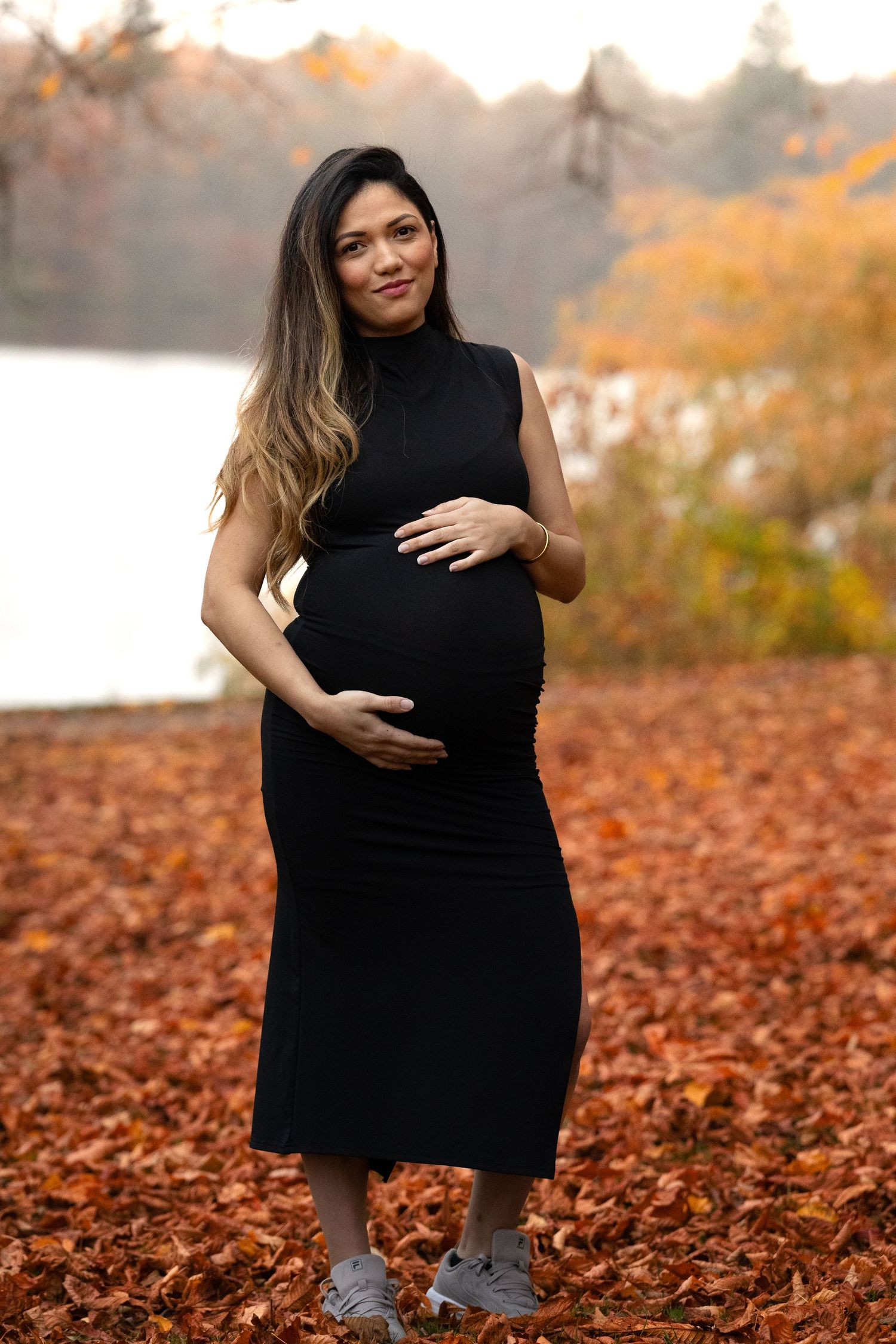 Smiling pregnant woman in black sleeveless dress holding baby bump surrounded by autumn foliage.