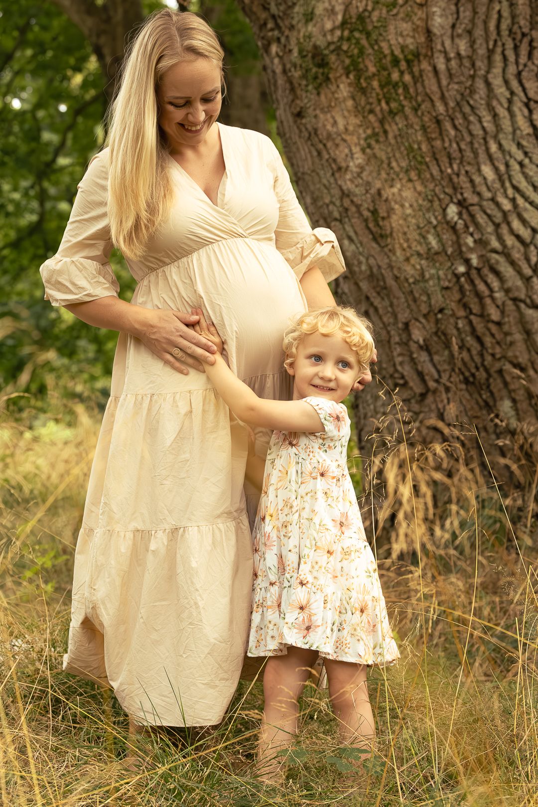 Pregnant blonde woman in cream dress with young daughter hugging her baby bump outdoors.