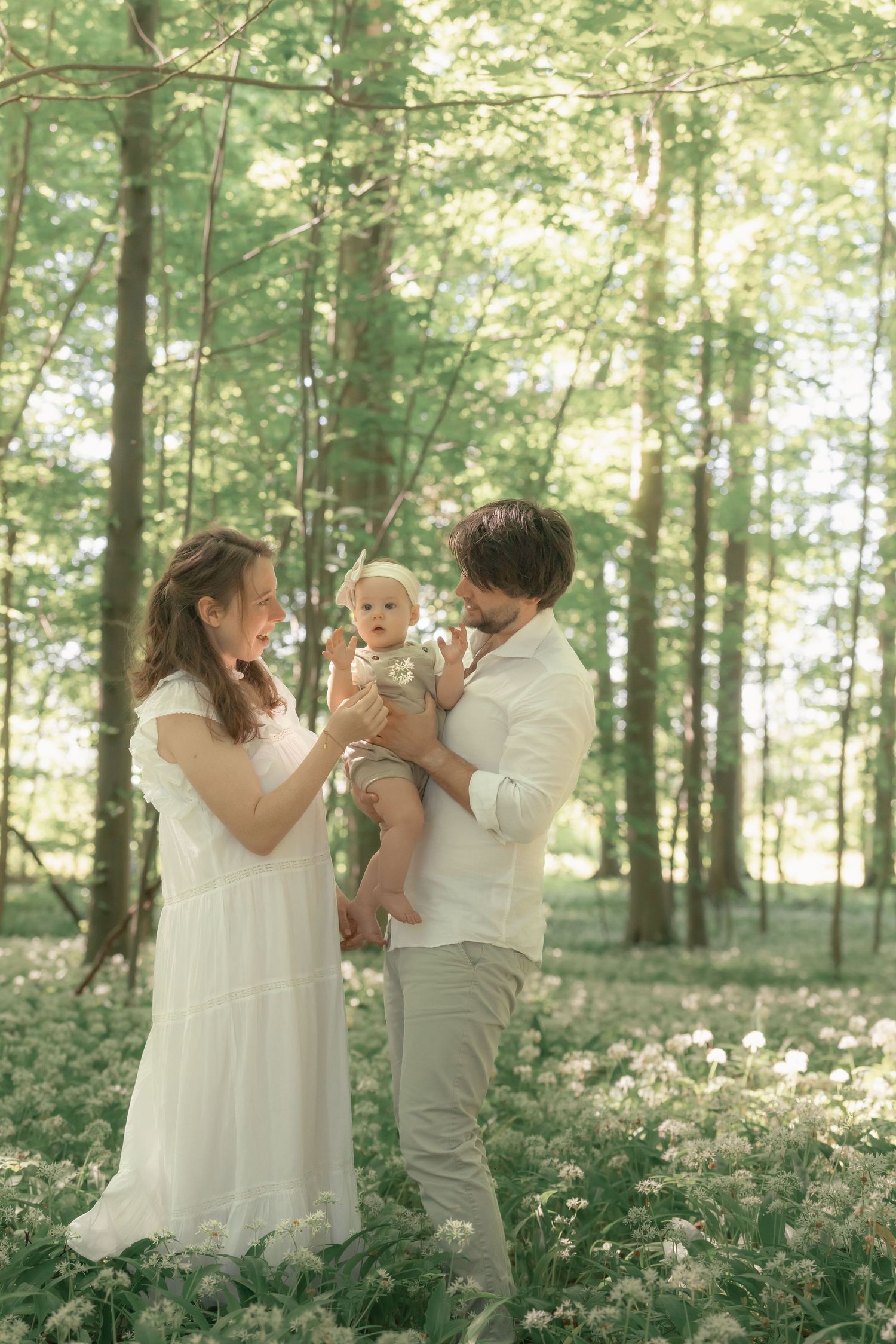 Happy young family in white outfits holding baby among wildflowers in green forest.