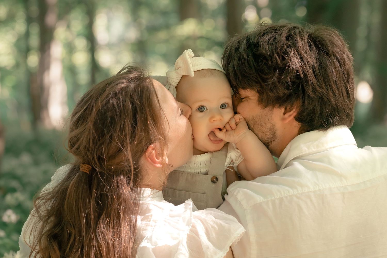 Loving parents simultaneously kissing their baby girl on cheeks in sunlit forest setting.