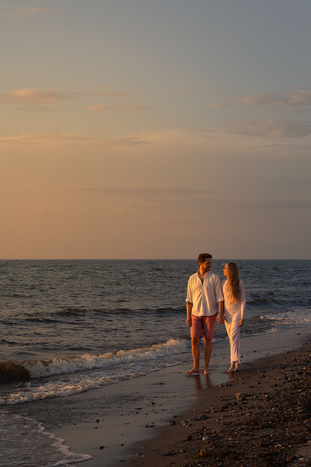 Romantic couple holding hands walking along shoreline at sunset with ocean waves in background.