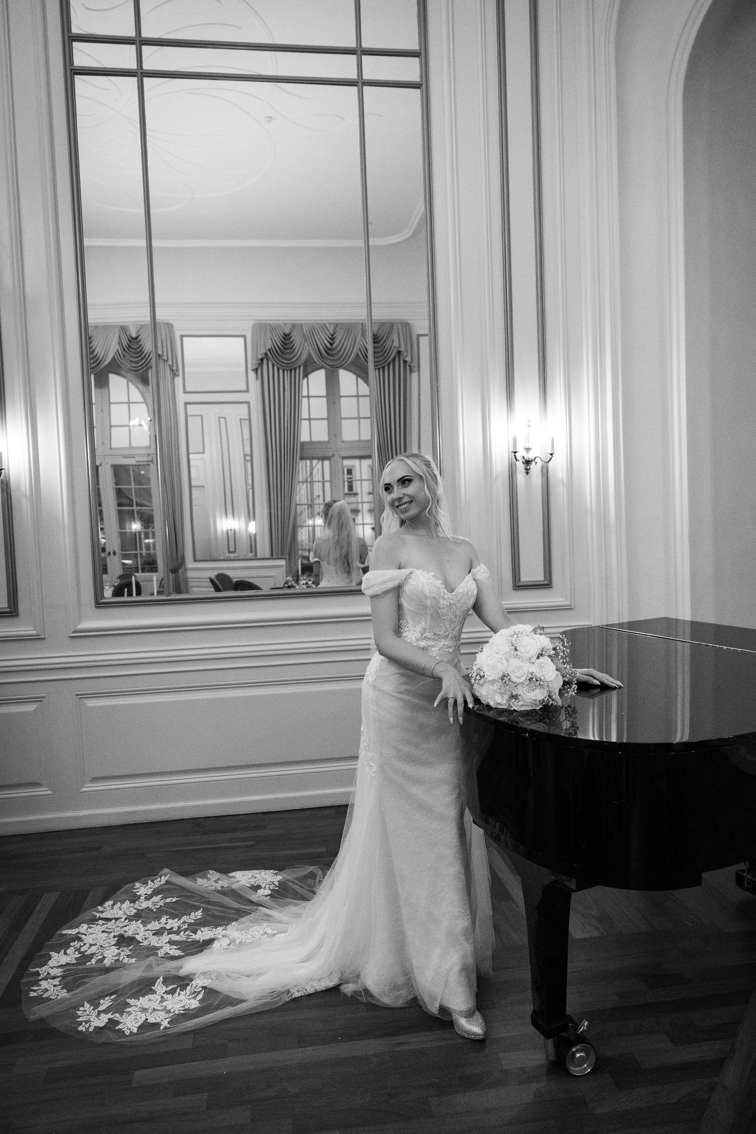 Elegant bride in lace gown poses beside grand piano holding bouquet in black and white portrait.