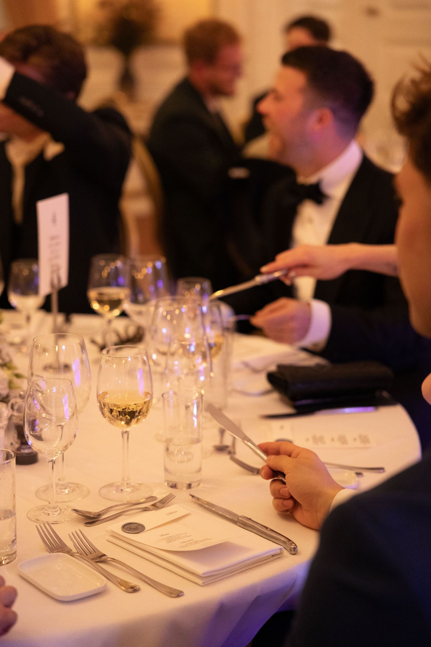 Guests in formal attire seated at elegantly set wedding reception dinner tables with wine glasses.