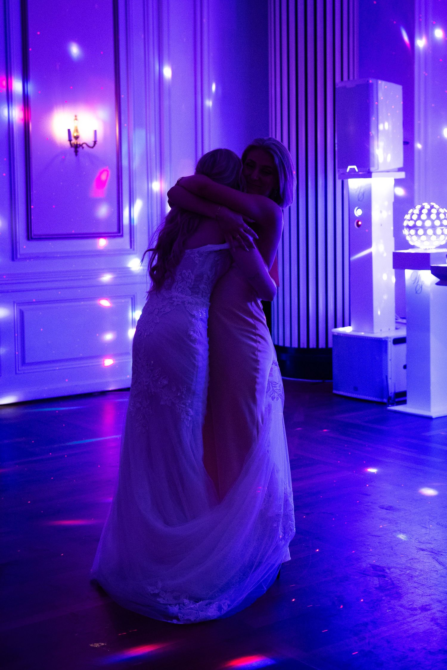 Two women in white lace gowns share a warm embrace on a dance floor under purple starry wedding reception lights.