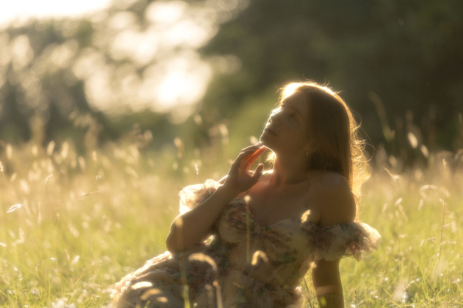 Woman in floral dress sitting in sunlit golden meadow, bathed in warm glowing backlight among tall grasses.
