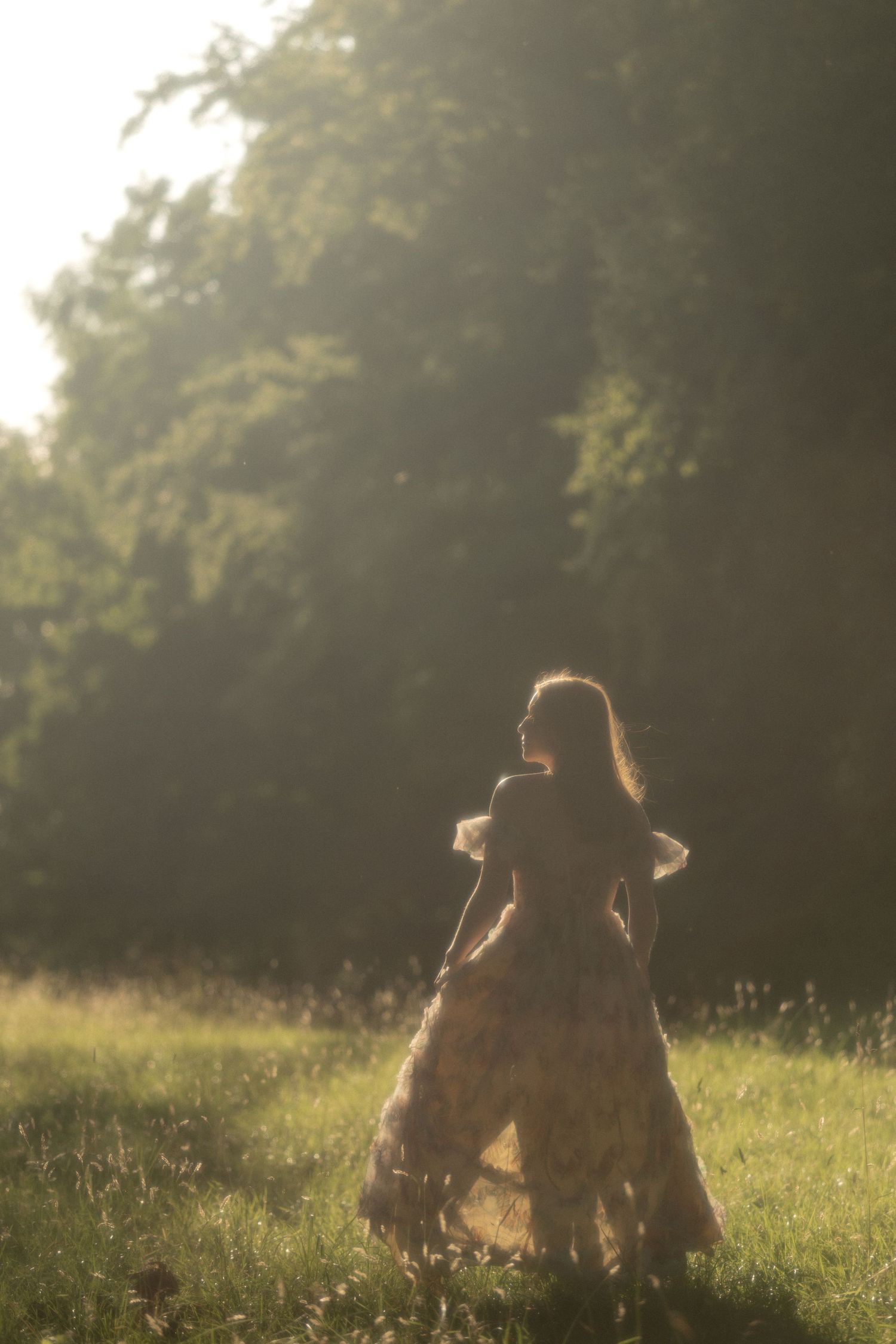 A woman in a flowing floral dress stands in a sunlit meadow surrounded by trees in golden hour light.