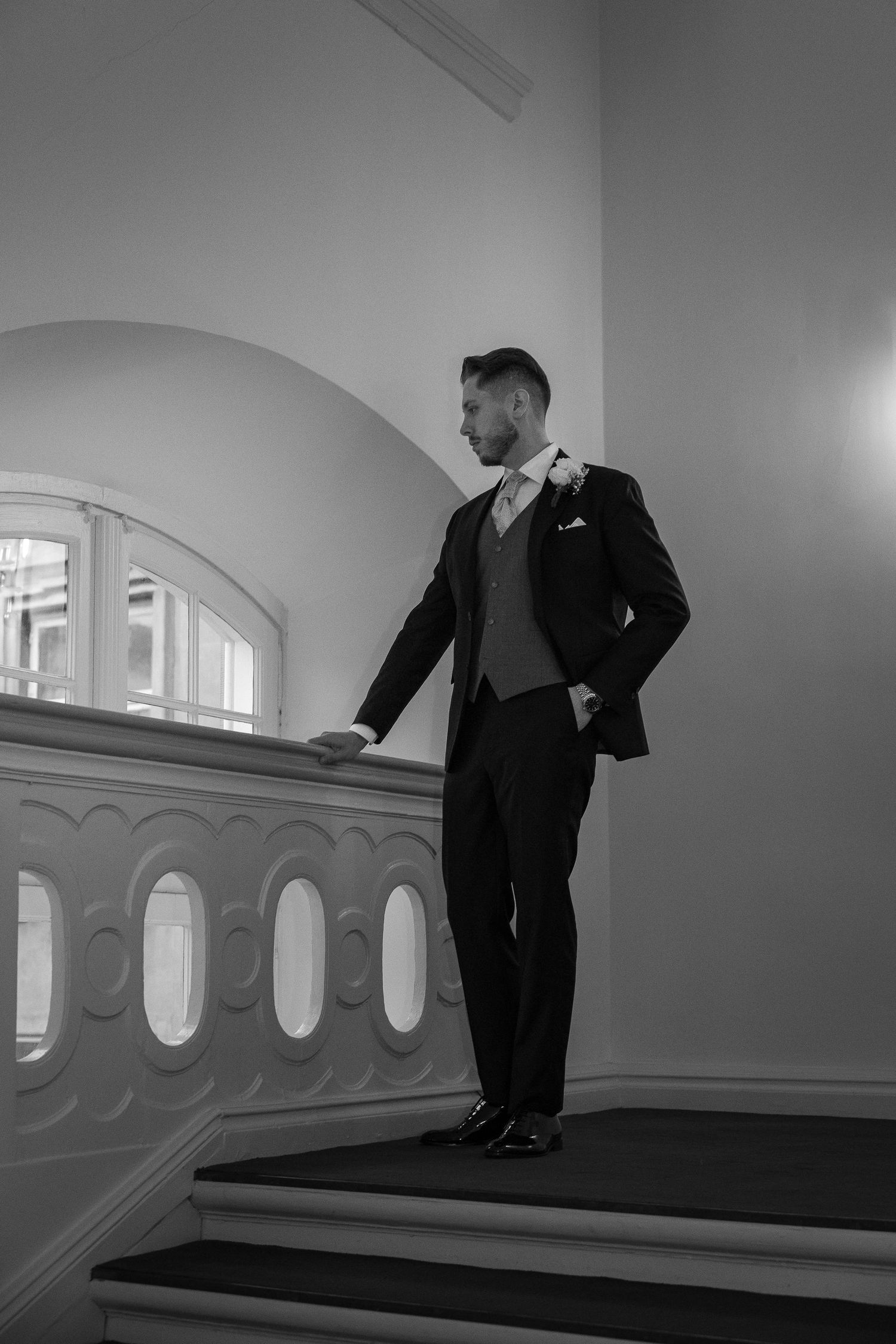Dashing groom in tailored suit poses on grand staircase balustrade in black and white portrait.