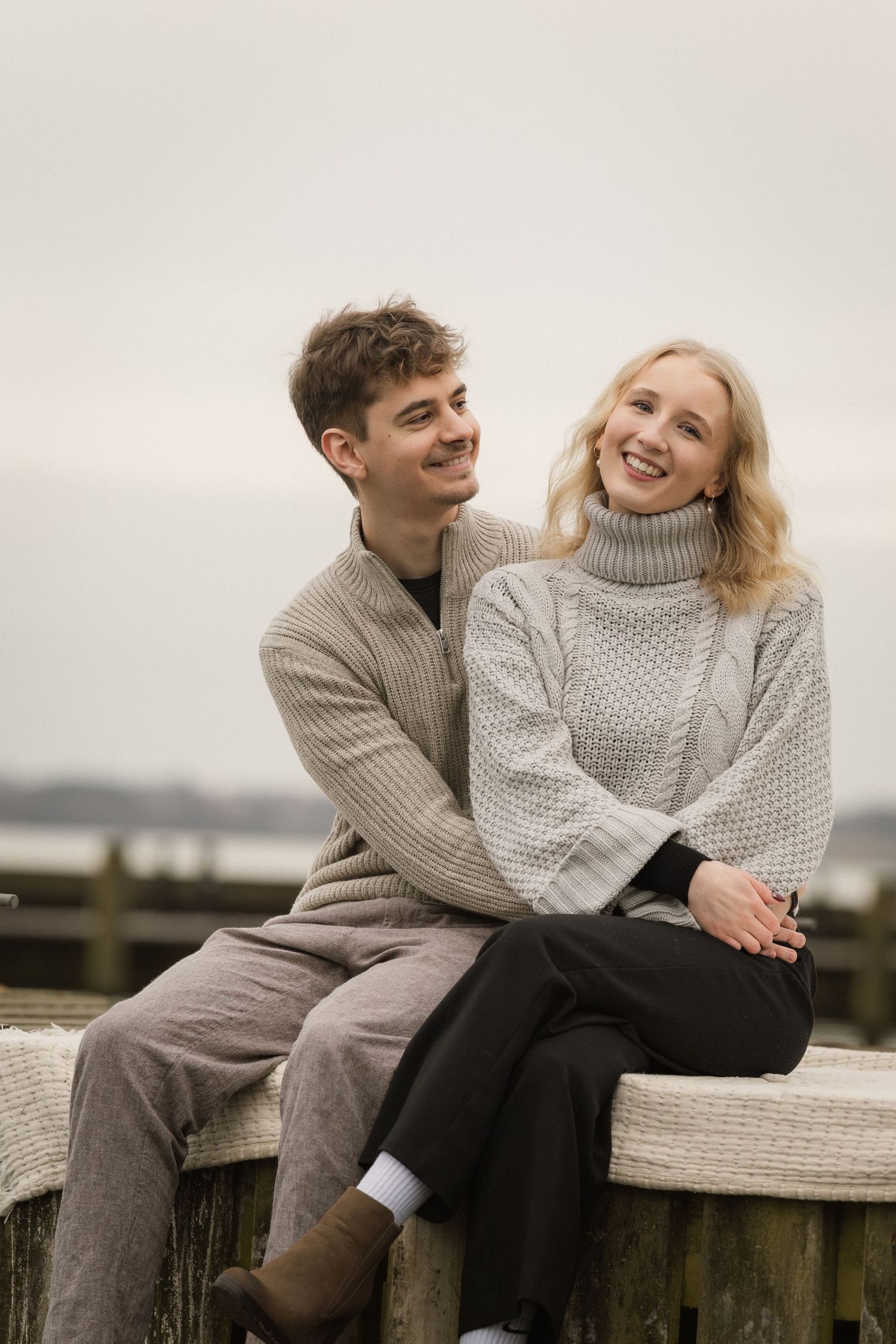 Smiling couple in cozy sweaters sitting on a waterfront dock on an overcast day.