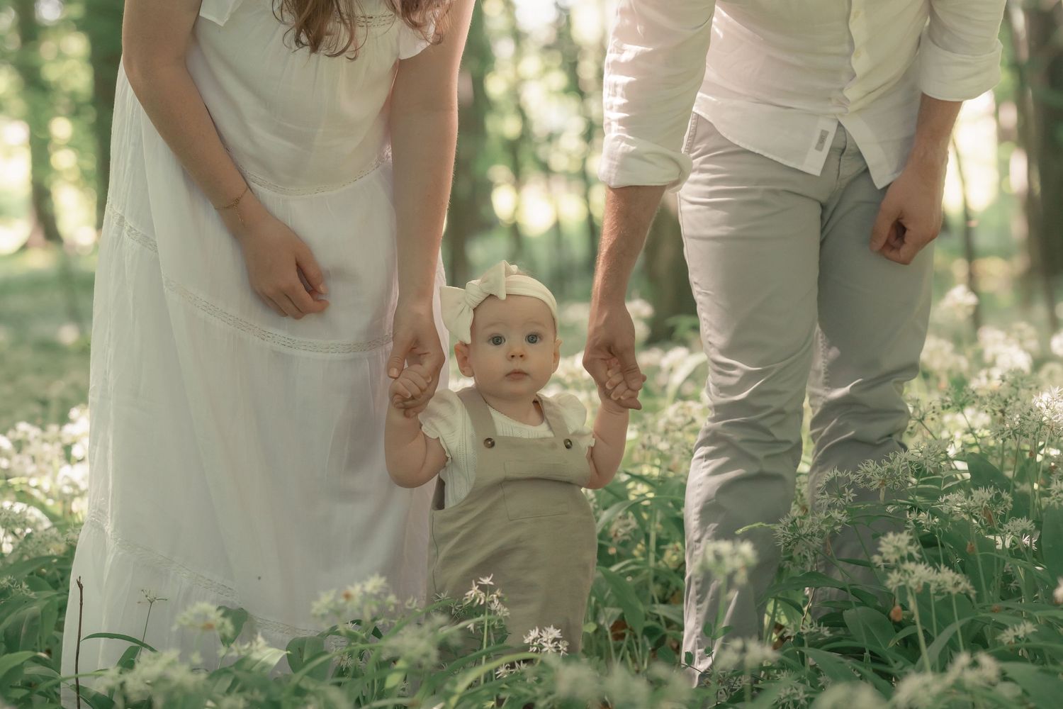 Baby takes first steps holding parents hands among white wildflowers in a green forest.