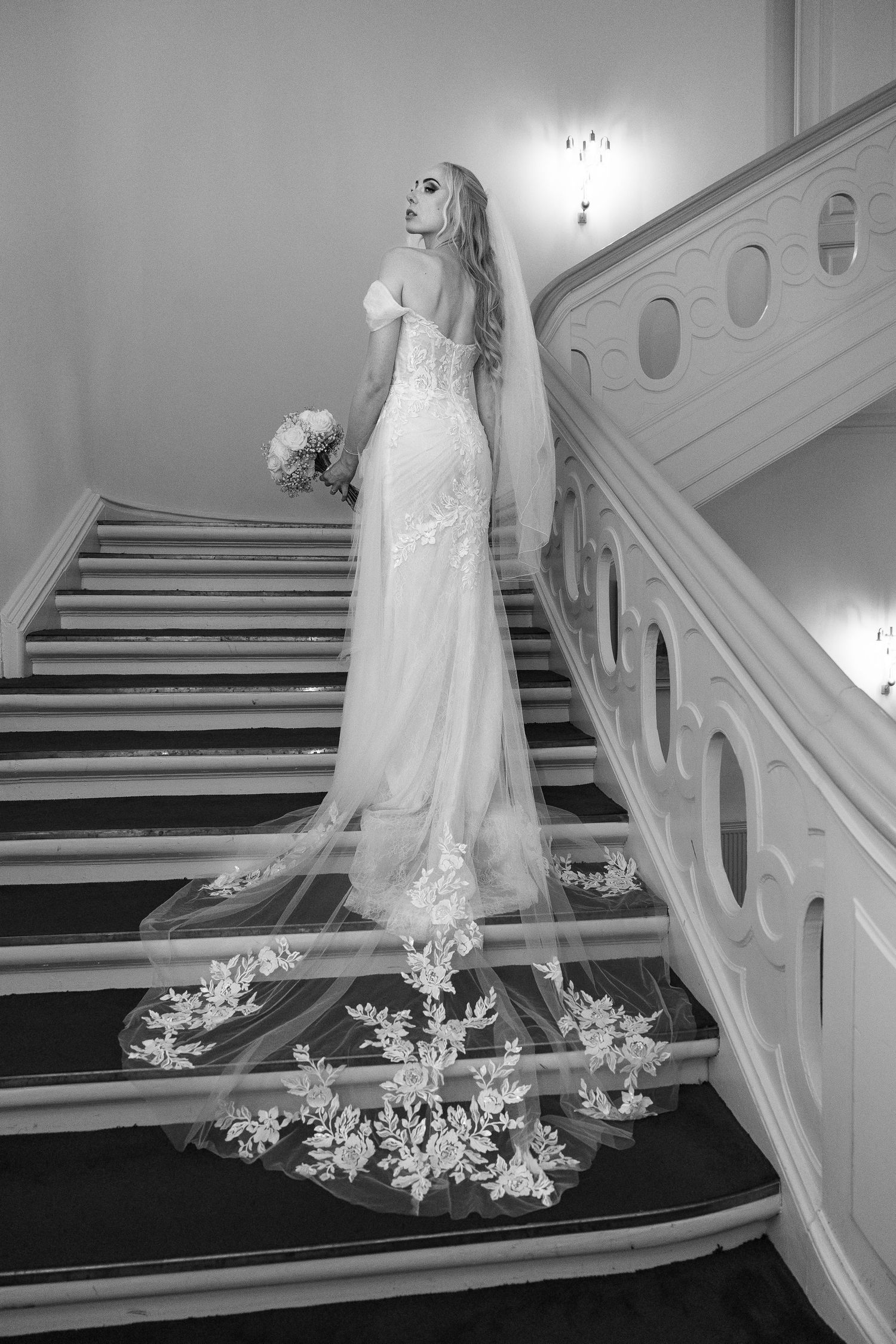 Bride in lace gown with floral cathedral veil posing on grand staircase.