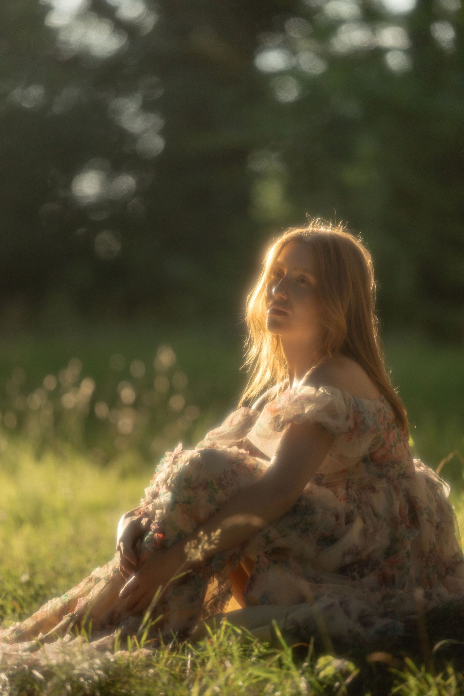 A woman in a floral dress sits on grass, bathed in golden sunset light with a soft bokeh green background.