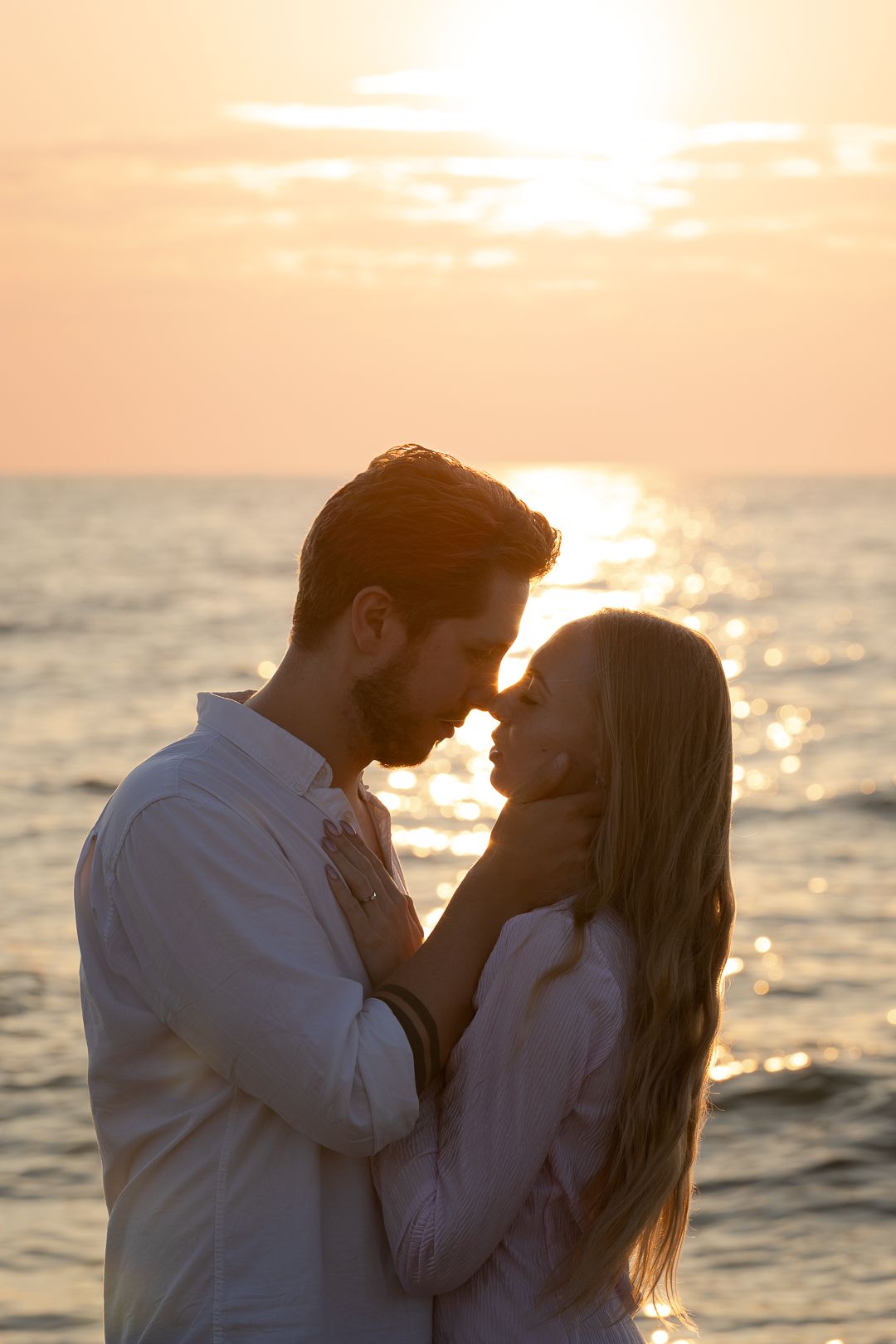A romantic couple nearly kissing in silhouette against a golden ocean sunset with warm glowing light.