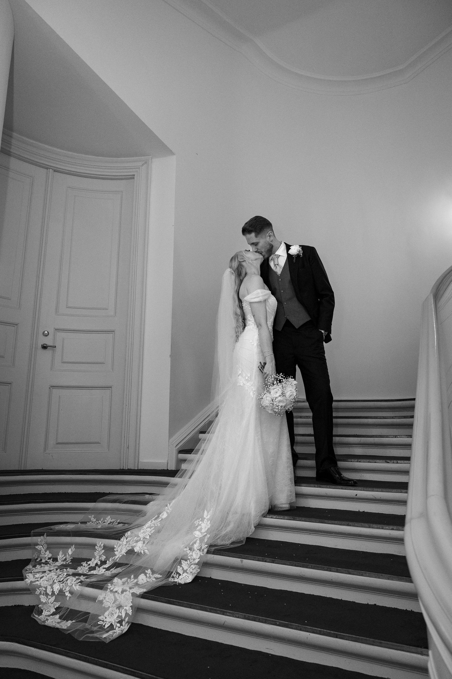 Bride and groom share romantic kiss on grand staircase, bridal veil trailing in black and white.