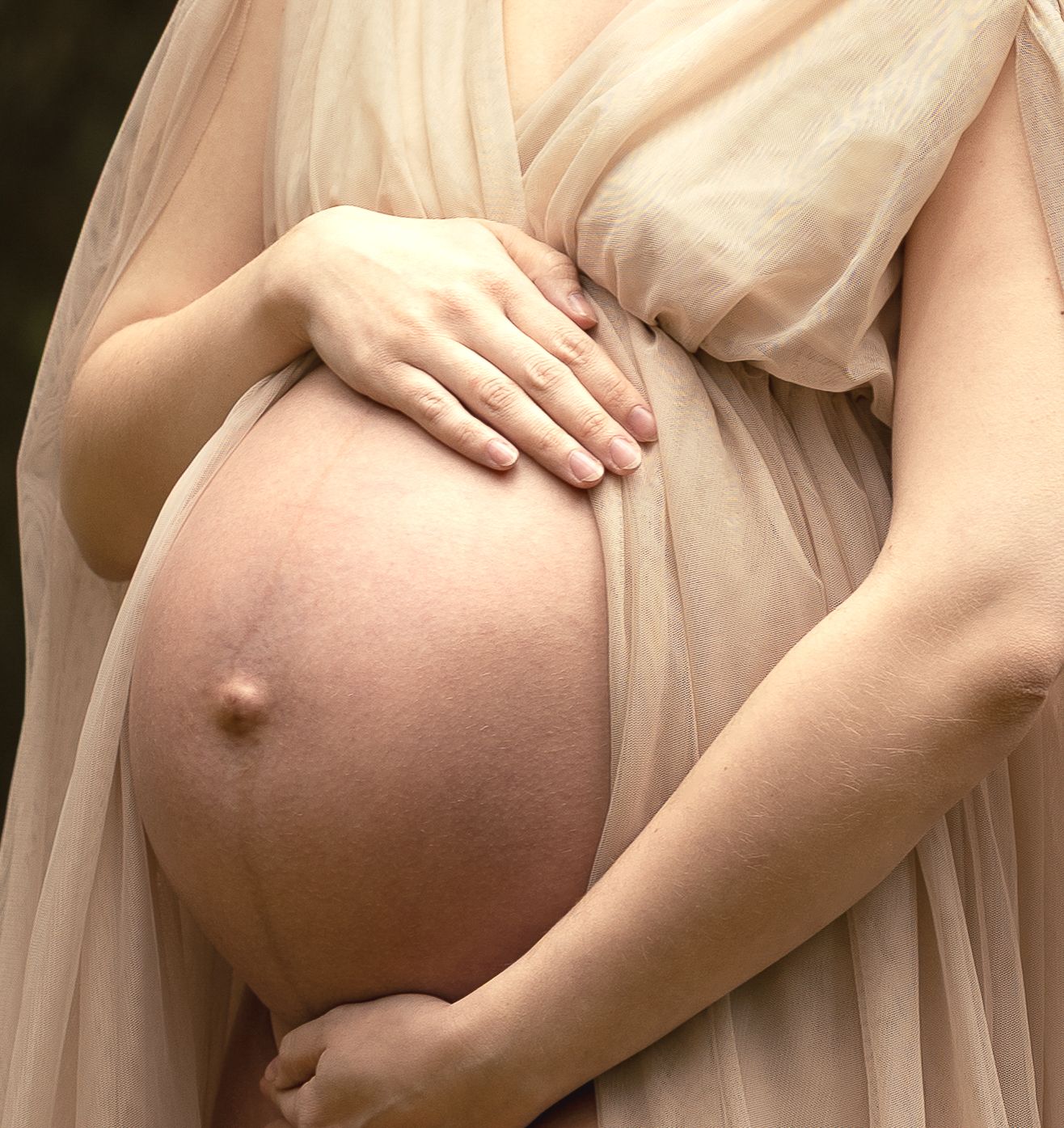 Close-up of pregnant woman in beige sheer dress cradling bare baby bump with both hands.