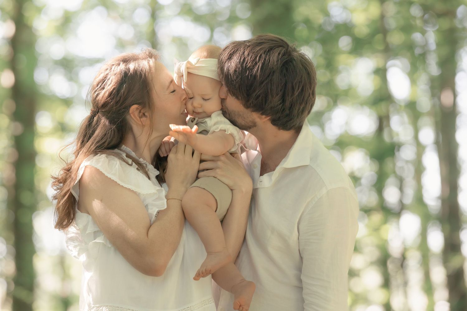 Happy parents share a tender moment kissing their smiling baby in a sunlit forest.