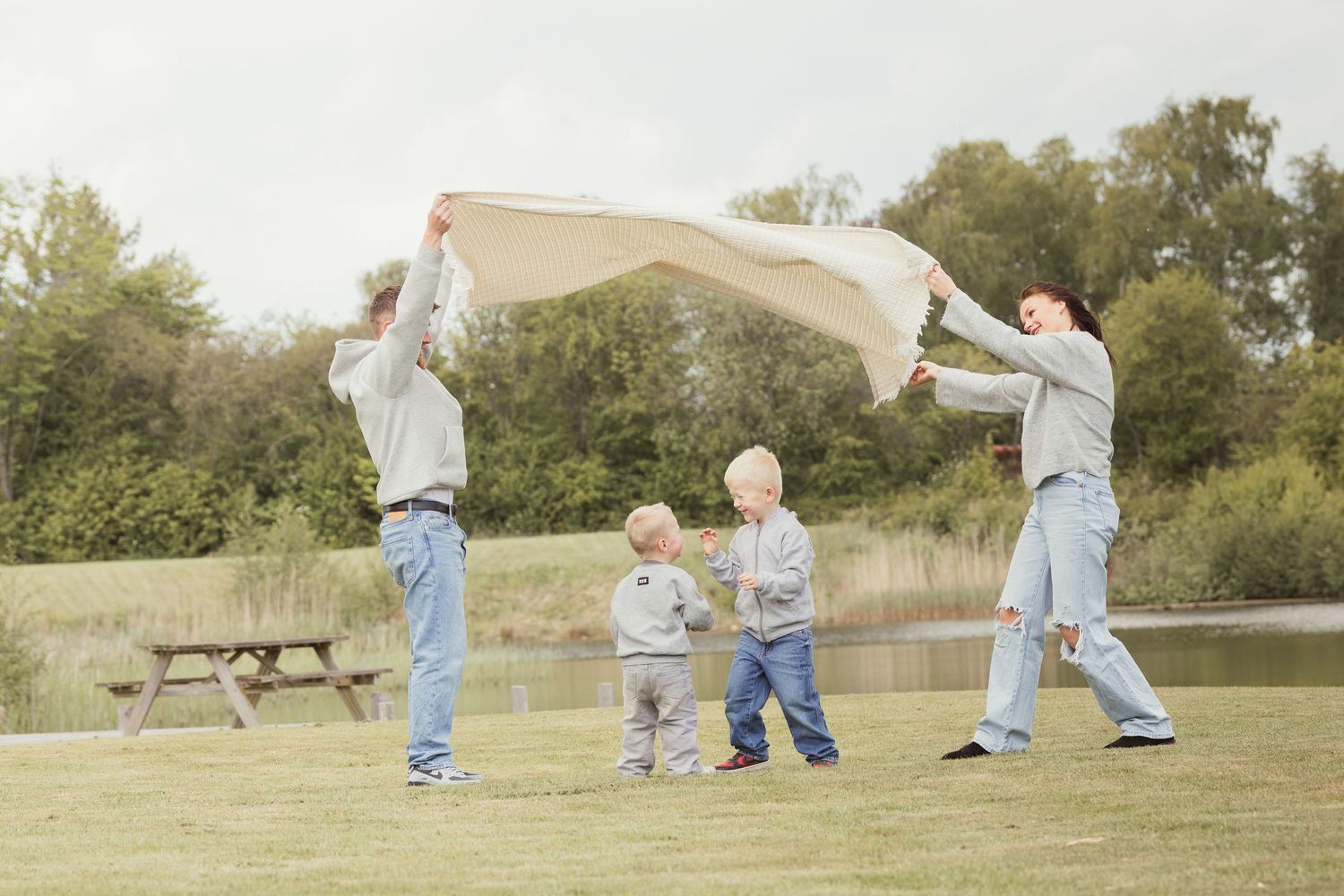 Family of four playing with blanket outdoors near pond in matching grey and blue outfits.